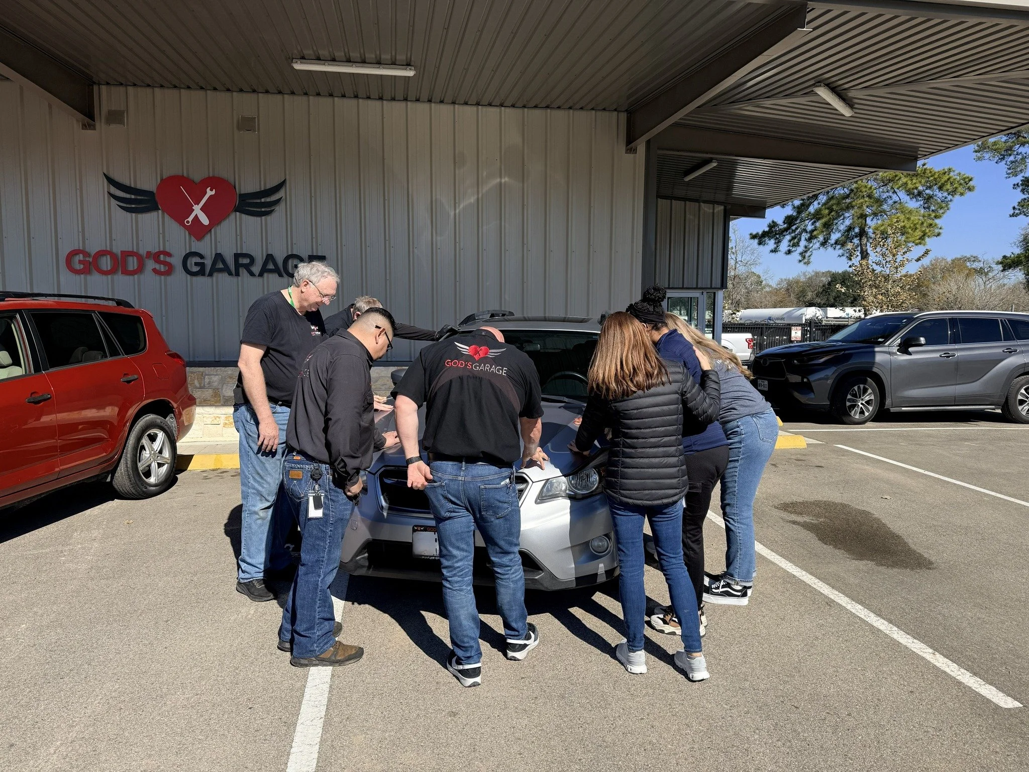 Group of people gathered around a car hood outside 'God's Garage' automotive shop.