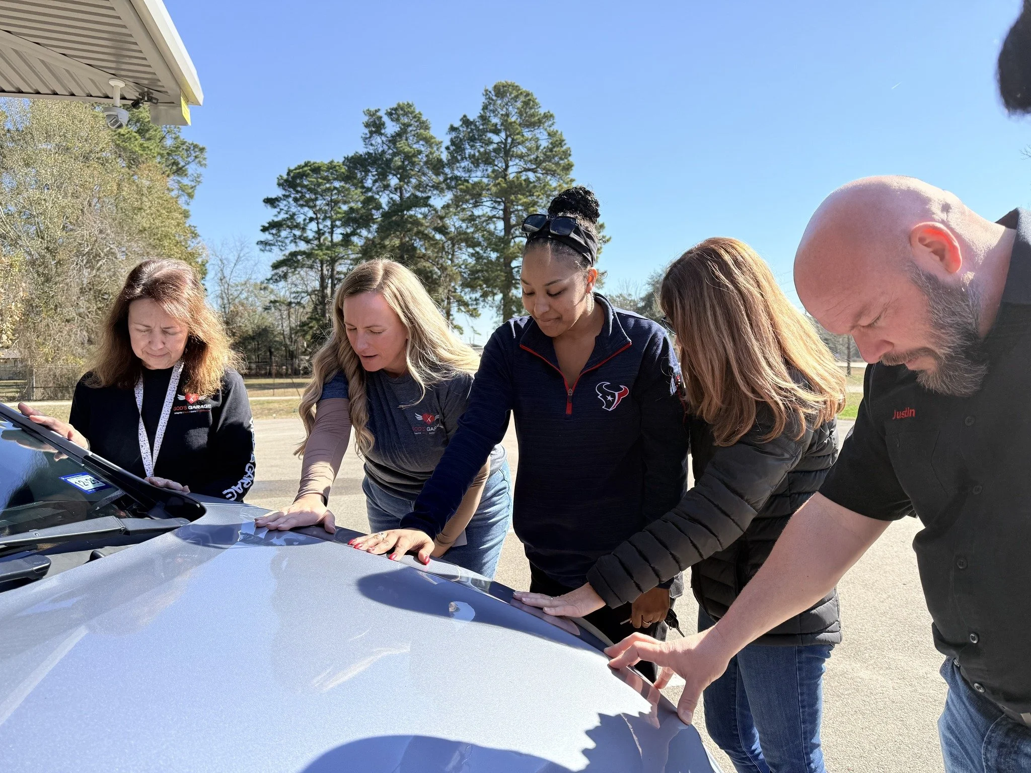 Group of five people inspecting a car hood outdoors on a sunny day.