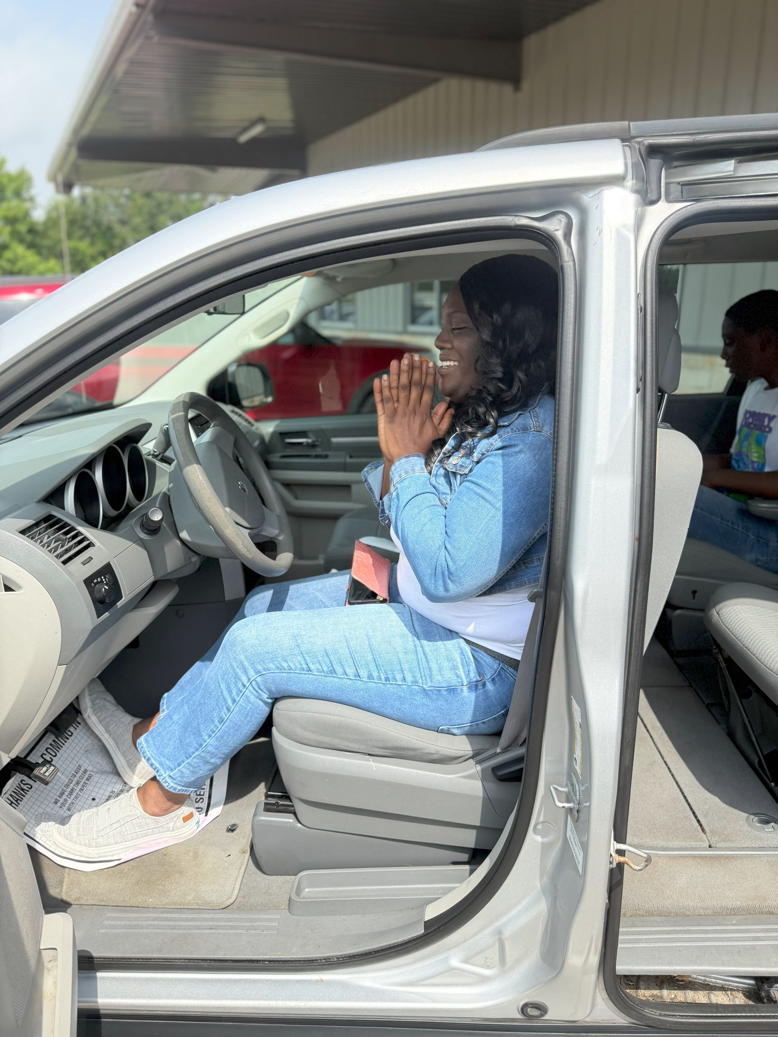 A woman sitting in the driver's seat of a silver van, smiling with hands clasped together, wearing a denim jacket and jeans, with a person in the back seat.