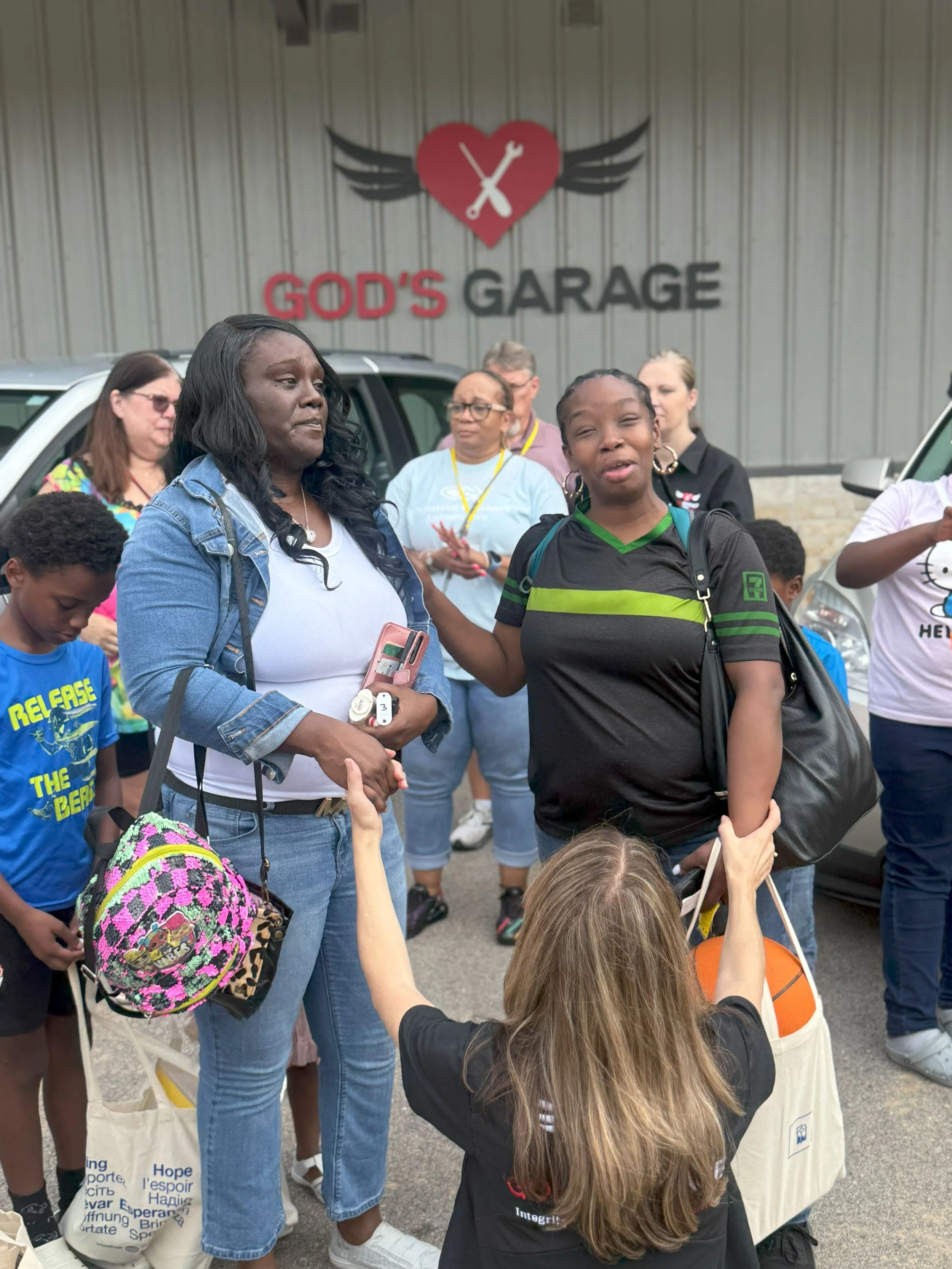 A group of people gathered outside a building with a sign that reads 'God's Garage.' Two women are in the foreground having a conversation, while others stand and sit around them. One woman has long hair, a denim jacket, and is holding a pink phone. 