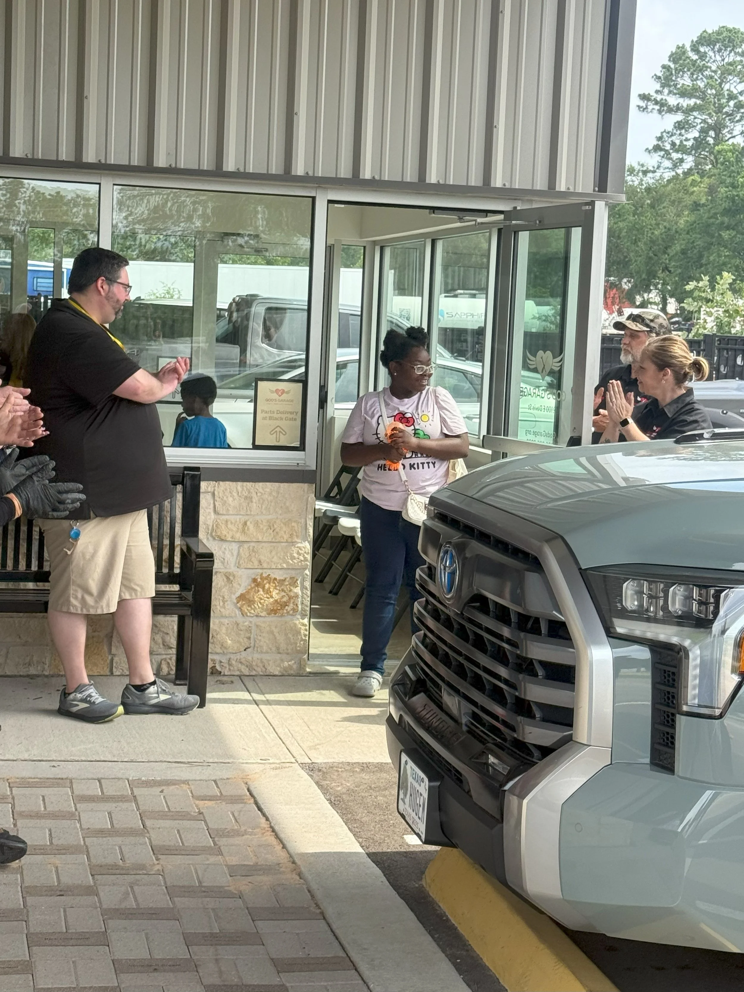 People standing outside a building, some clapping, with a woman in a Hello Kitty shirt near the entrance, and a gray pickup truck parked in front.