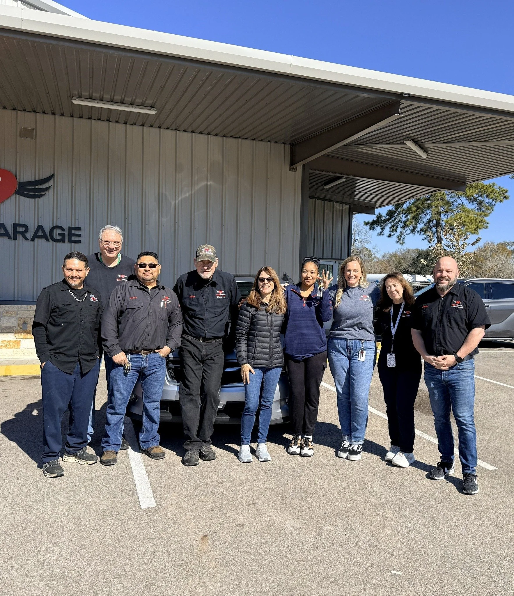Group of nine people standing in front of a building with a garage sign, smiling, on a sunny day.