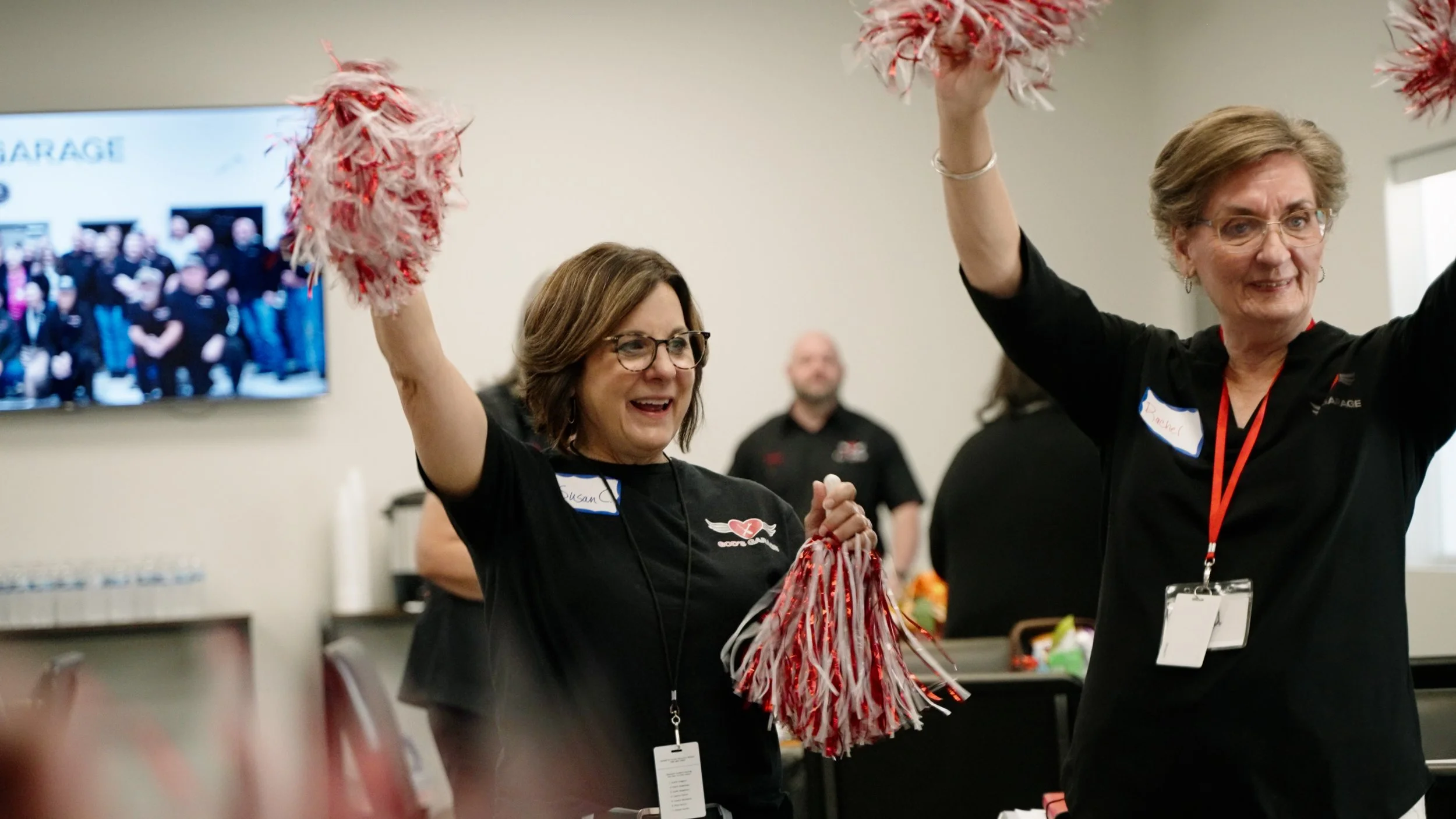 Two women at a celebration event holding red and white pom-poms, smiling and cheering. They are wearing black shirts with name tags and badges, inside a room with a table and a TV screen in the background.