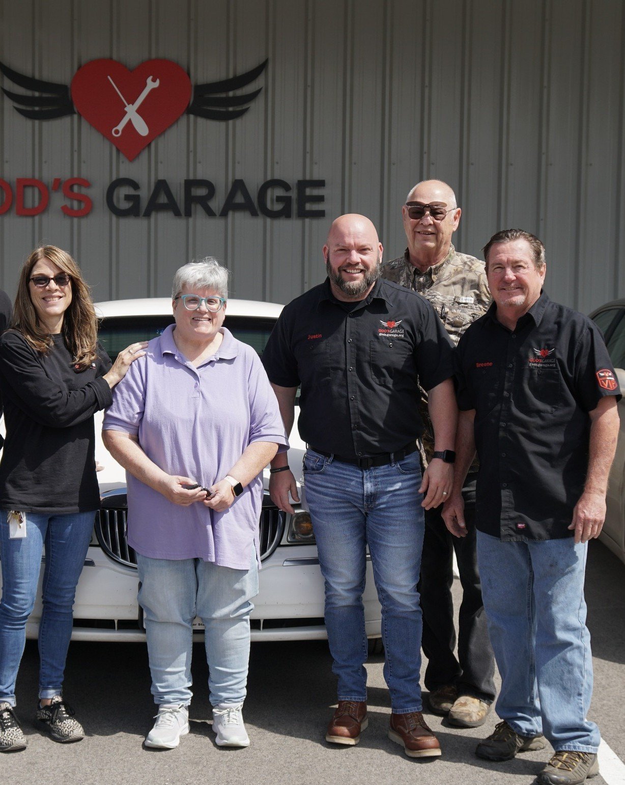 Group of five people standing outside a garage with a sign that reads "Todd's Garage" and a red heart with a wrench and screwdriver with wings on it.