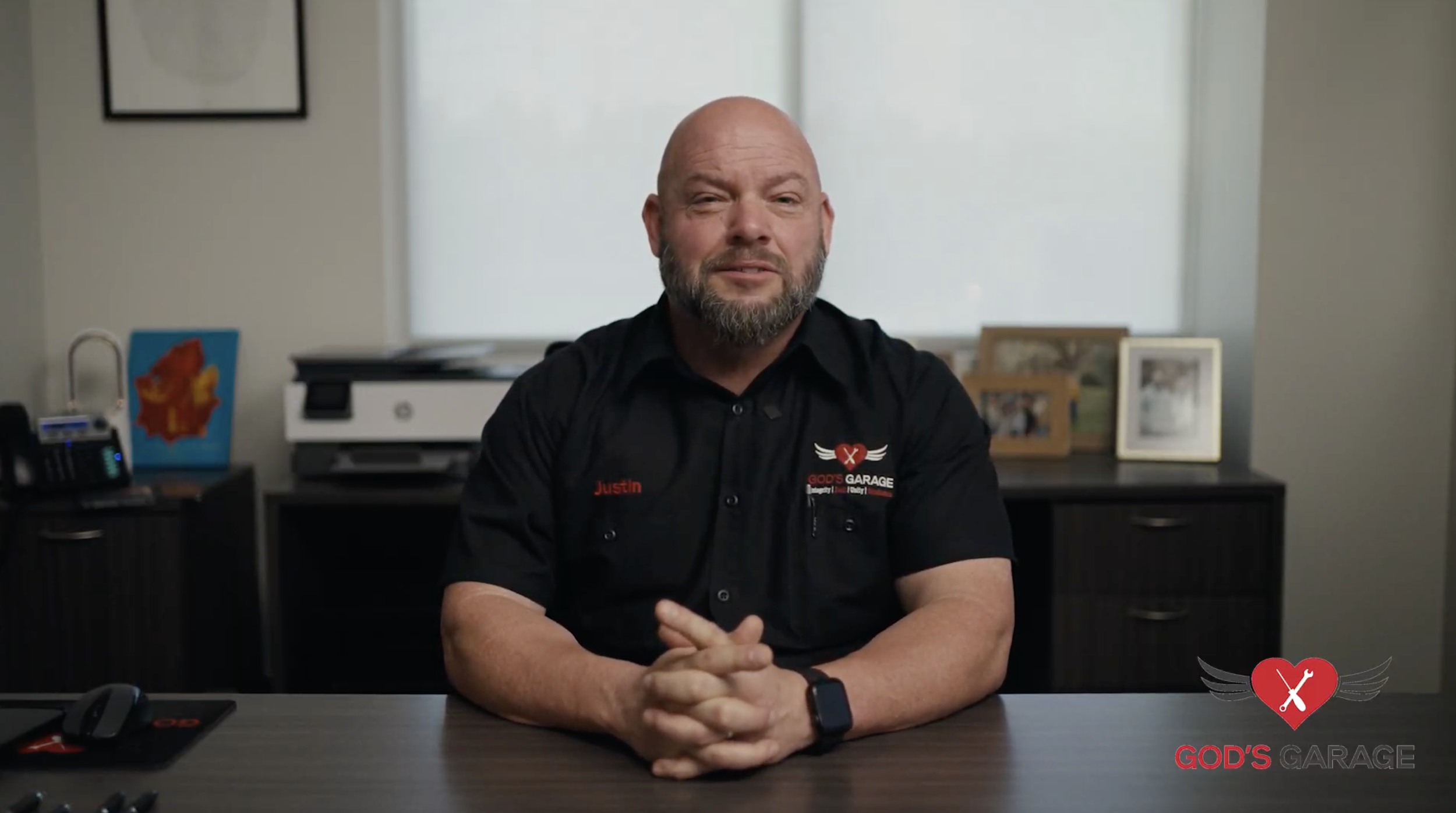 A man with a beard and bald head sitting at a desk in an office. He is wearing a black shirt with a logo and the name "Justin" embroidered on it. The background shows framed photos, a printer, and a window. The logo "GOD'S GARAGE" is visible in the bottom right corner of the image.