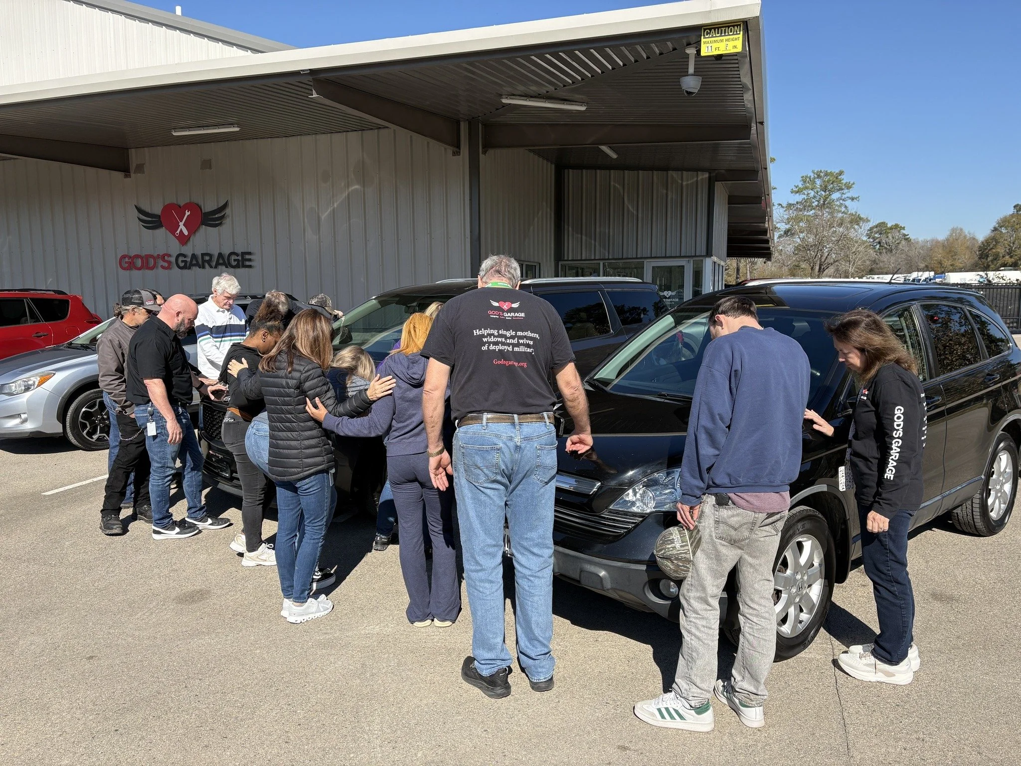Group of people prayerfully gathered around a black car in front of God's Garage, a community service organization, with a building and other cars in the background on a sunny day.