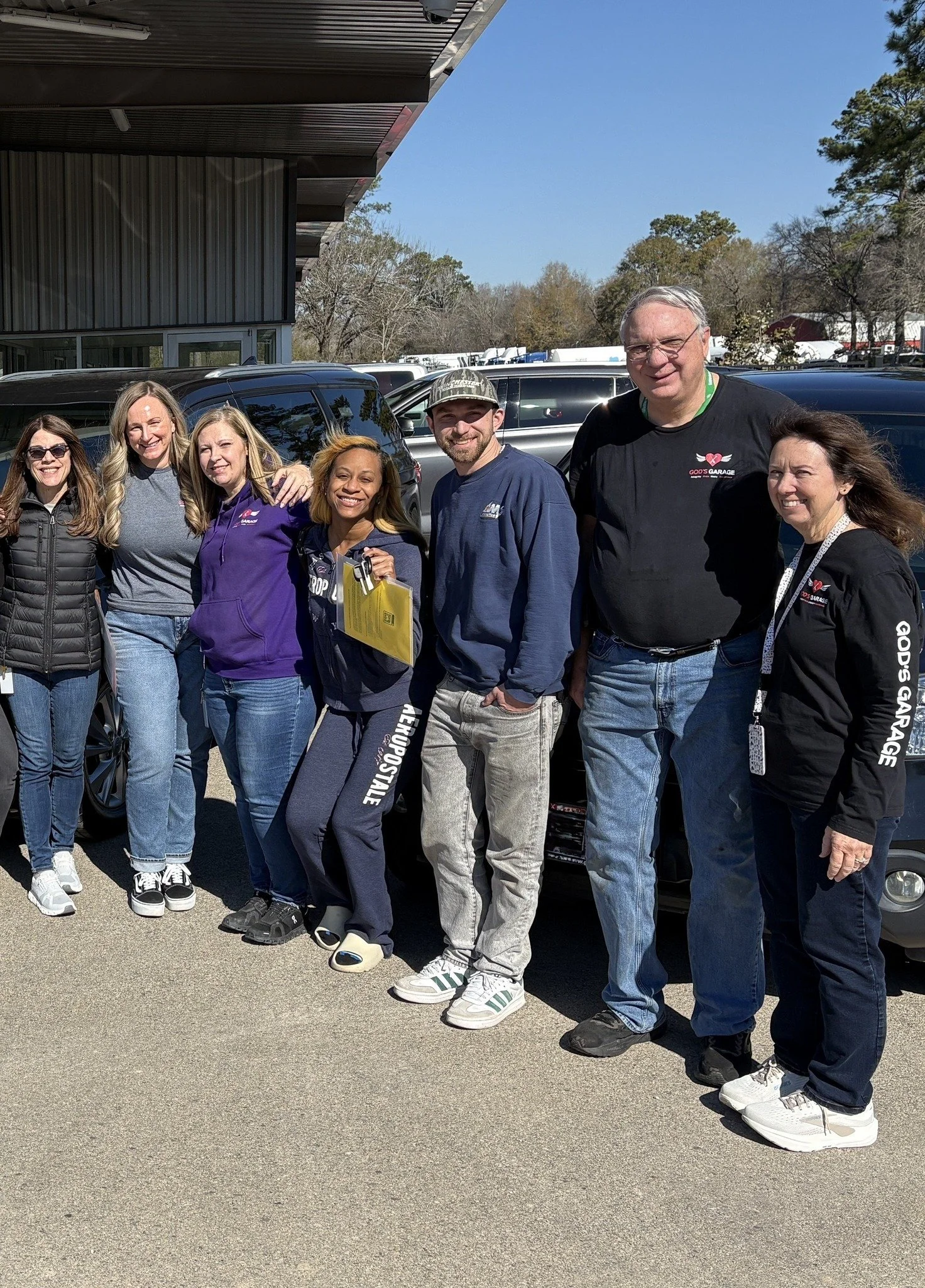 Group of seven people standing outdoors in front of parked cars, smiling for the photo.