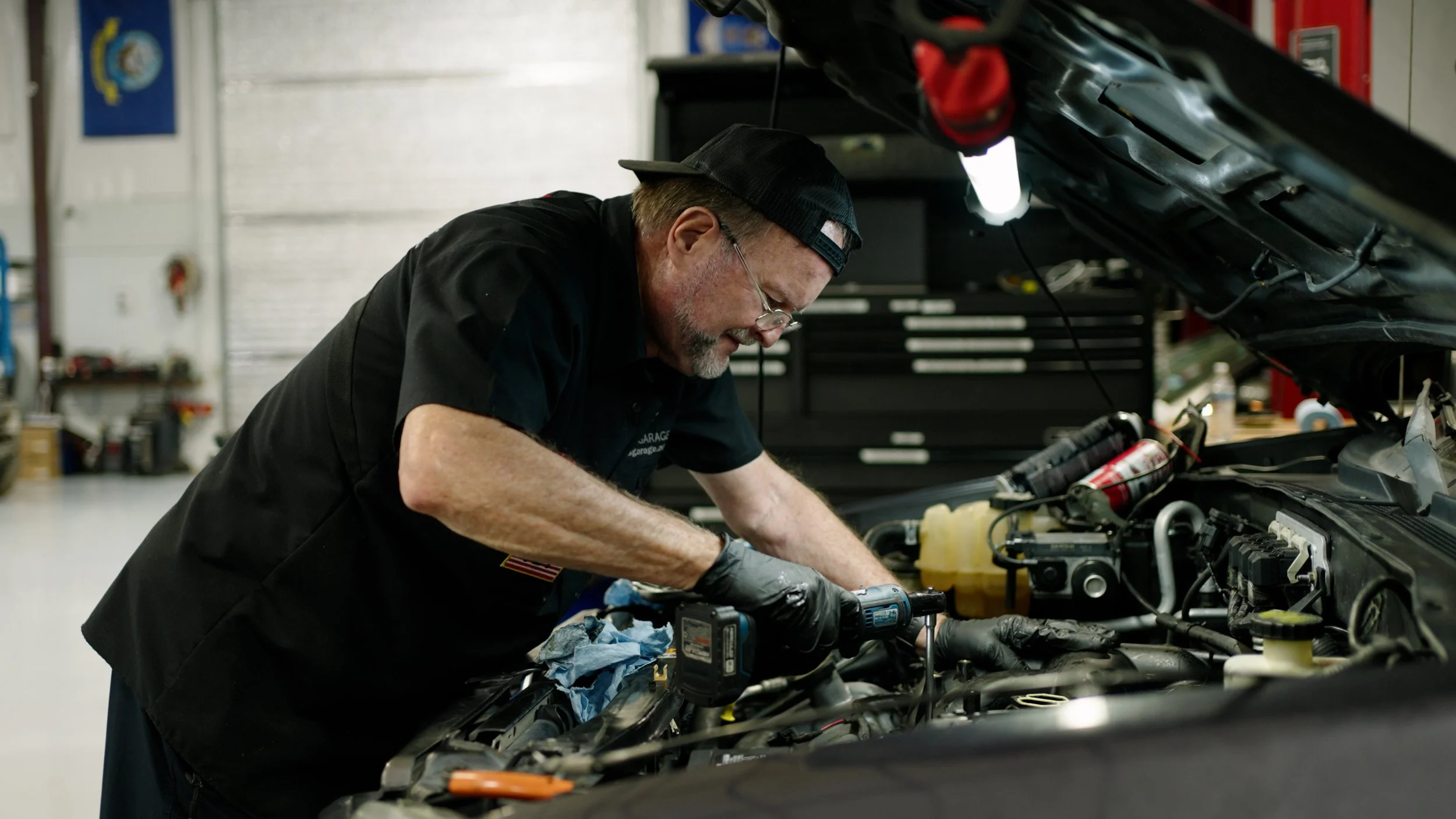 A mechanic working under the hood of a car in a garage, using a power tool to repair the engine.