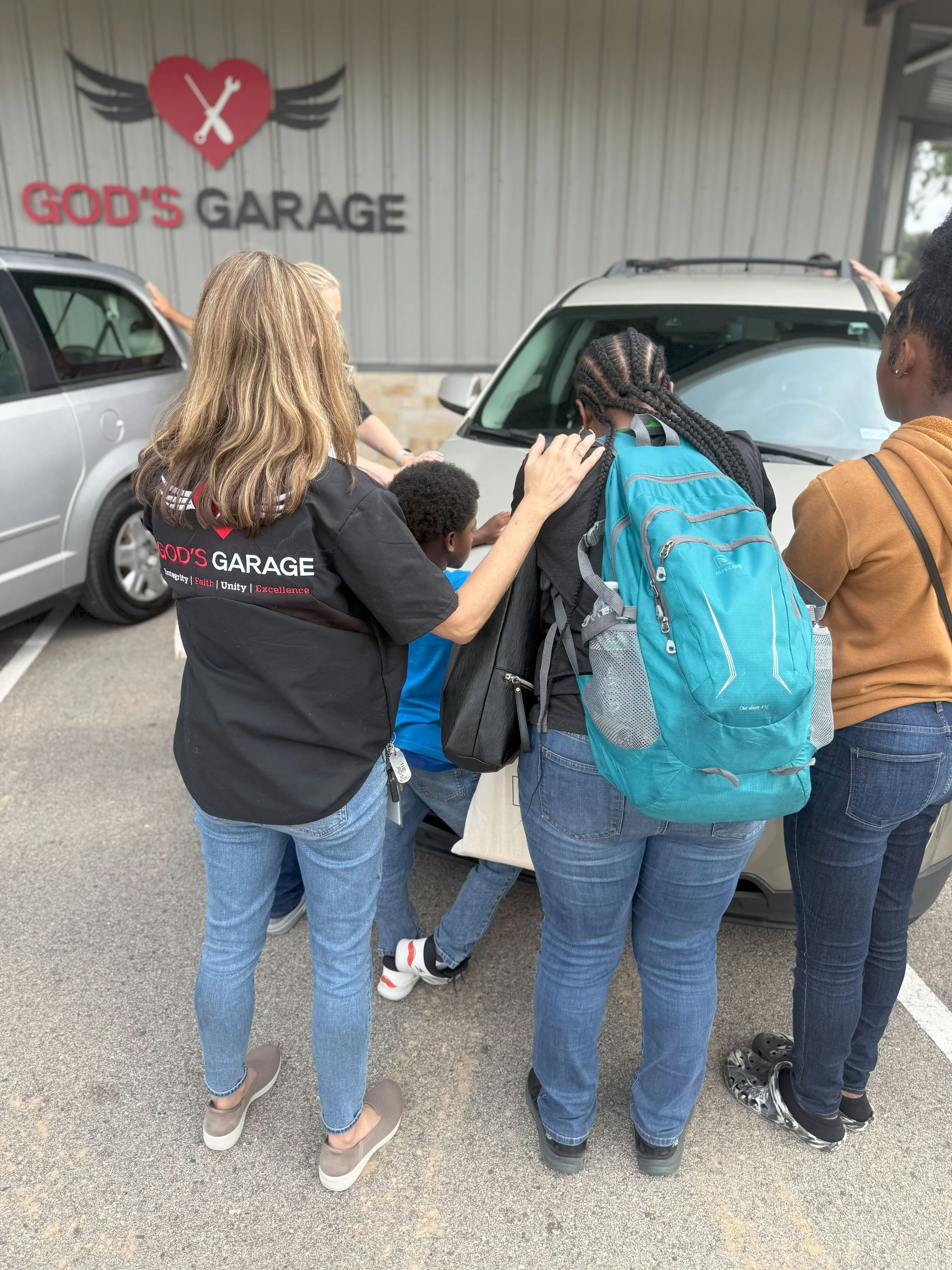 A group of people gathered outside a church named God's Garage, praying over a child leaning against a car, with a church sign featuring a heart with a wrench and wings in the background.