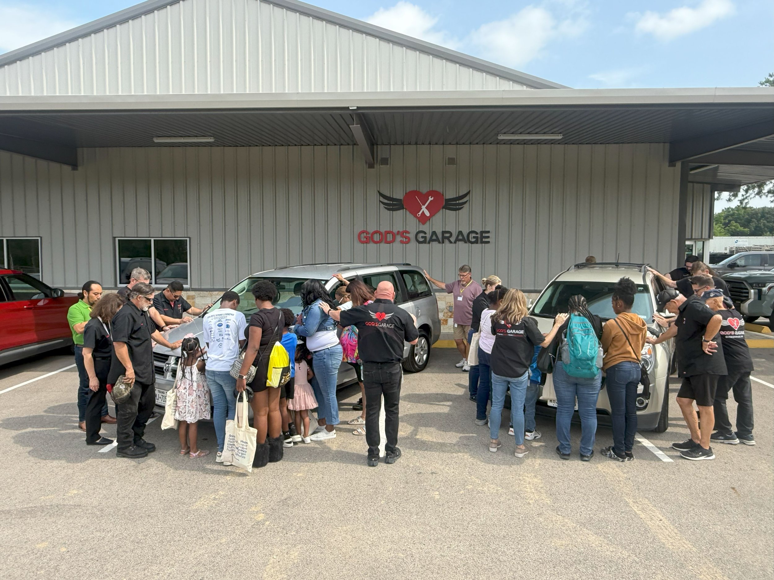 A group of people, including children, gathered in front of a building labeled 'GOD'S GARAGE,' with some people praying or touching cars parked outside.
