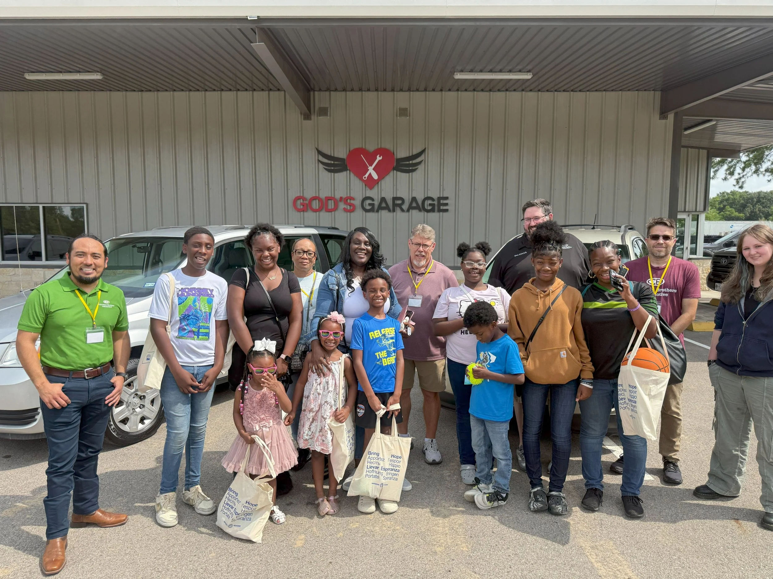 Group of people standing in front of a building with a sign that reads 'God's Garage,' some holding bags, posing for a photo on a parking lot.