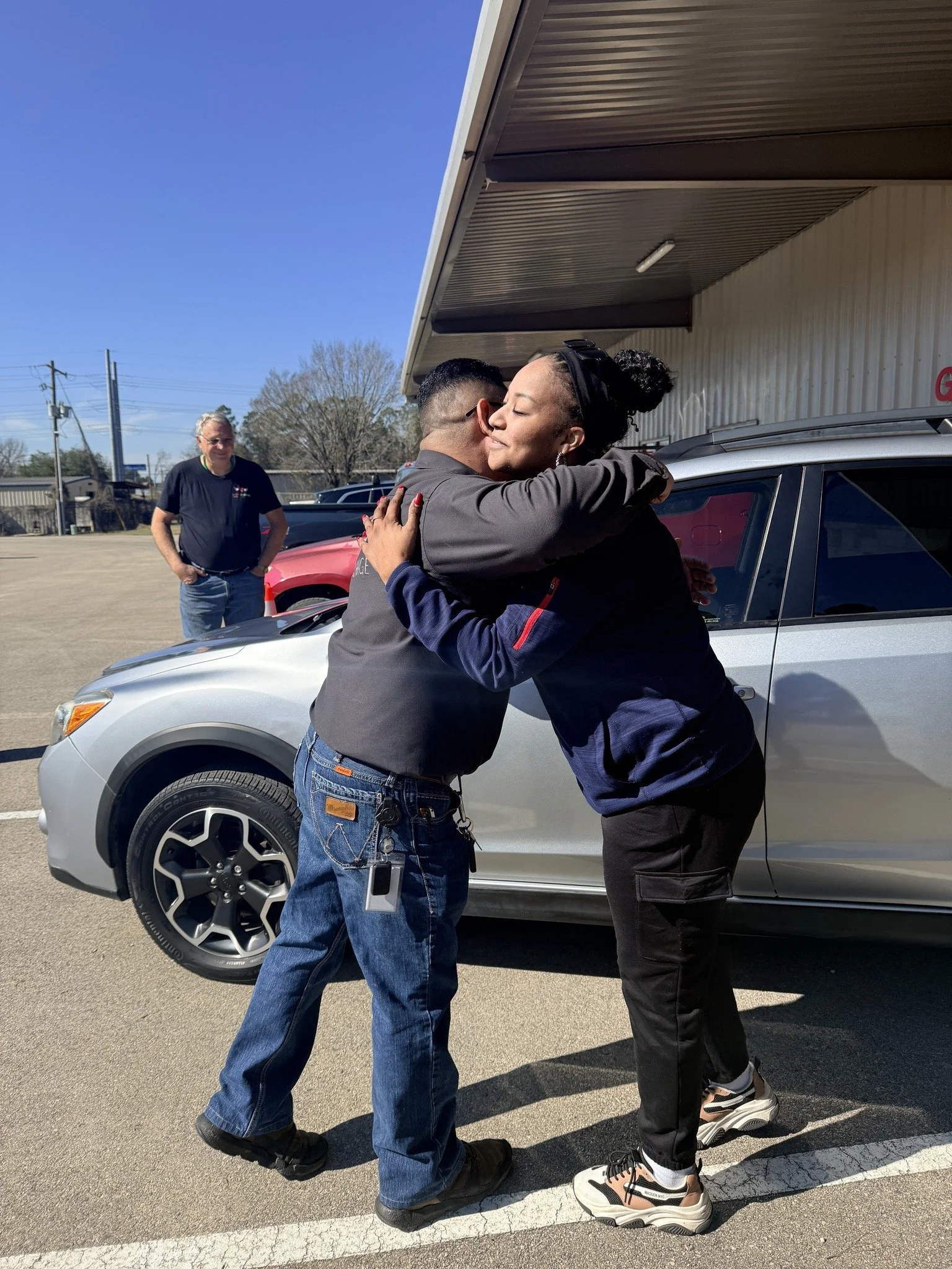 Two people hugging outside a building, a man and a woman, with a gray car behind them. A third person stands nearby, observing.