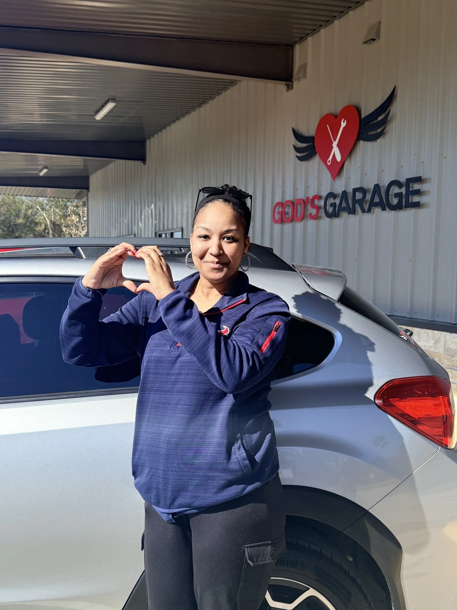 A woman wearing a blue jacket making a heart shape with her hands, standing in front of a silver car outside a building with a sign reading "God's Garage" and a logo of a fork and wrench with wings.