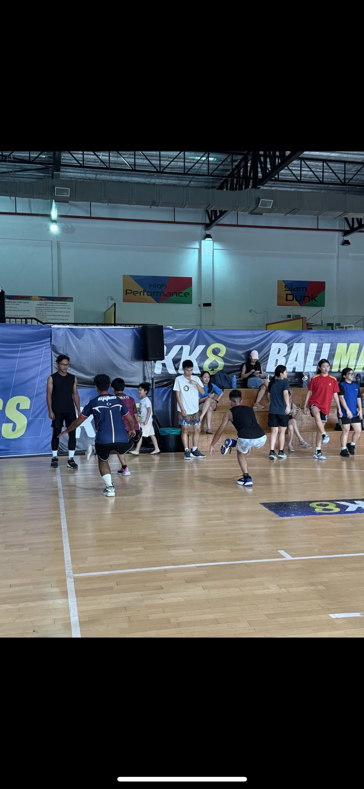 Kids playing volleyball in an indoor gymnasium with banners that read "Kicks & Slam Dunk" and people sitting and standing along the sidelines.