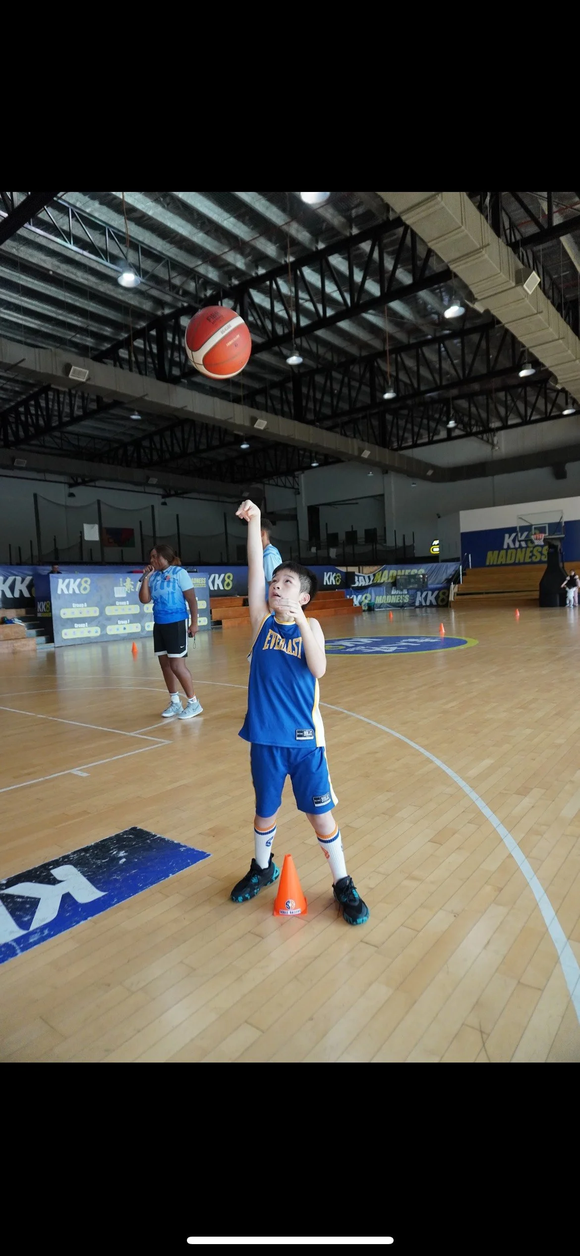 Young boy in blue basketball uniform practicing free throw shot on indoor basketball court, with basketball in mid-air and orange cone markers nearby.