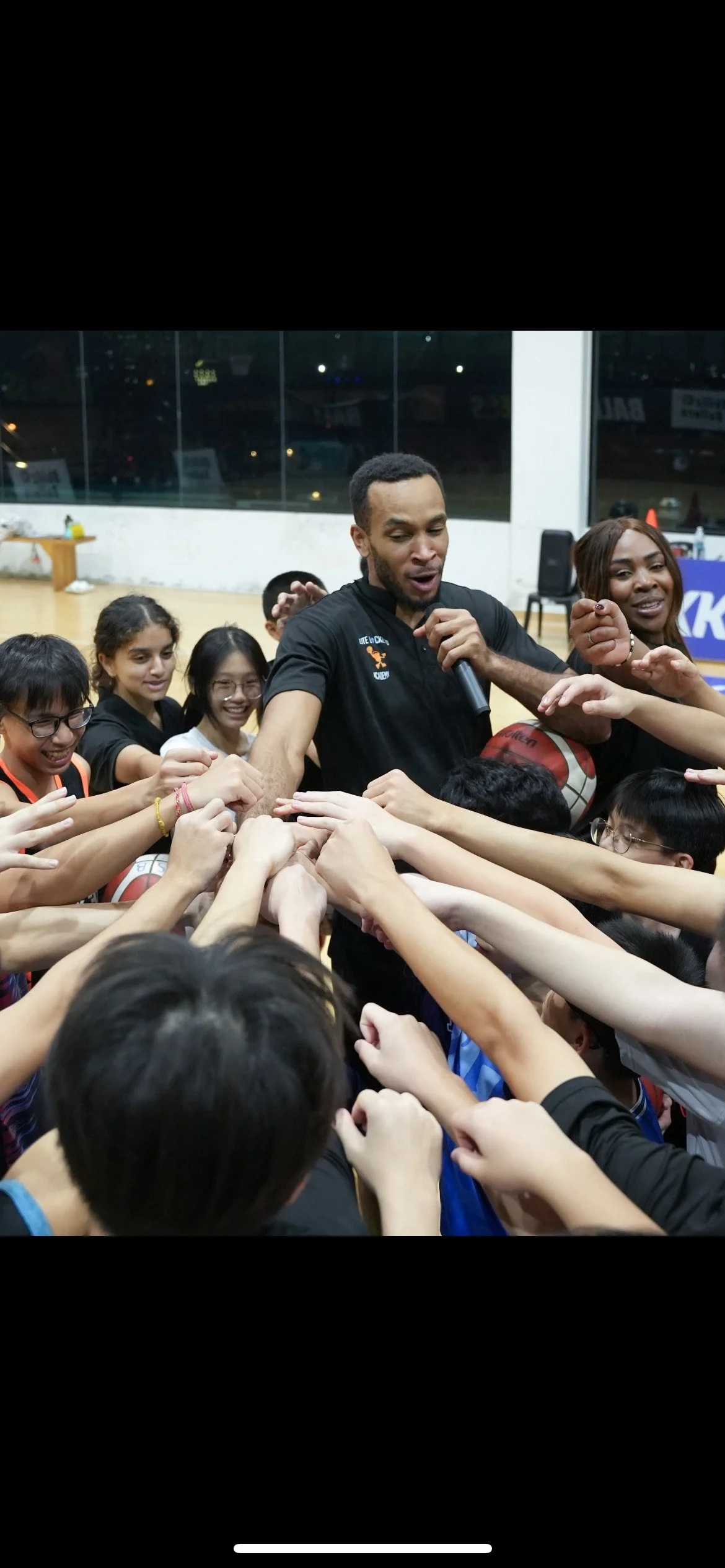 A man holding a microphone is surrounded by young girls and boys reaching out their hands towards him, creating a huddle during a basketball event in a gymnasium.