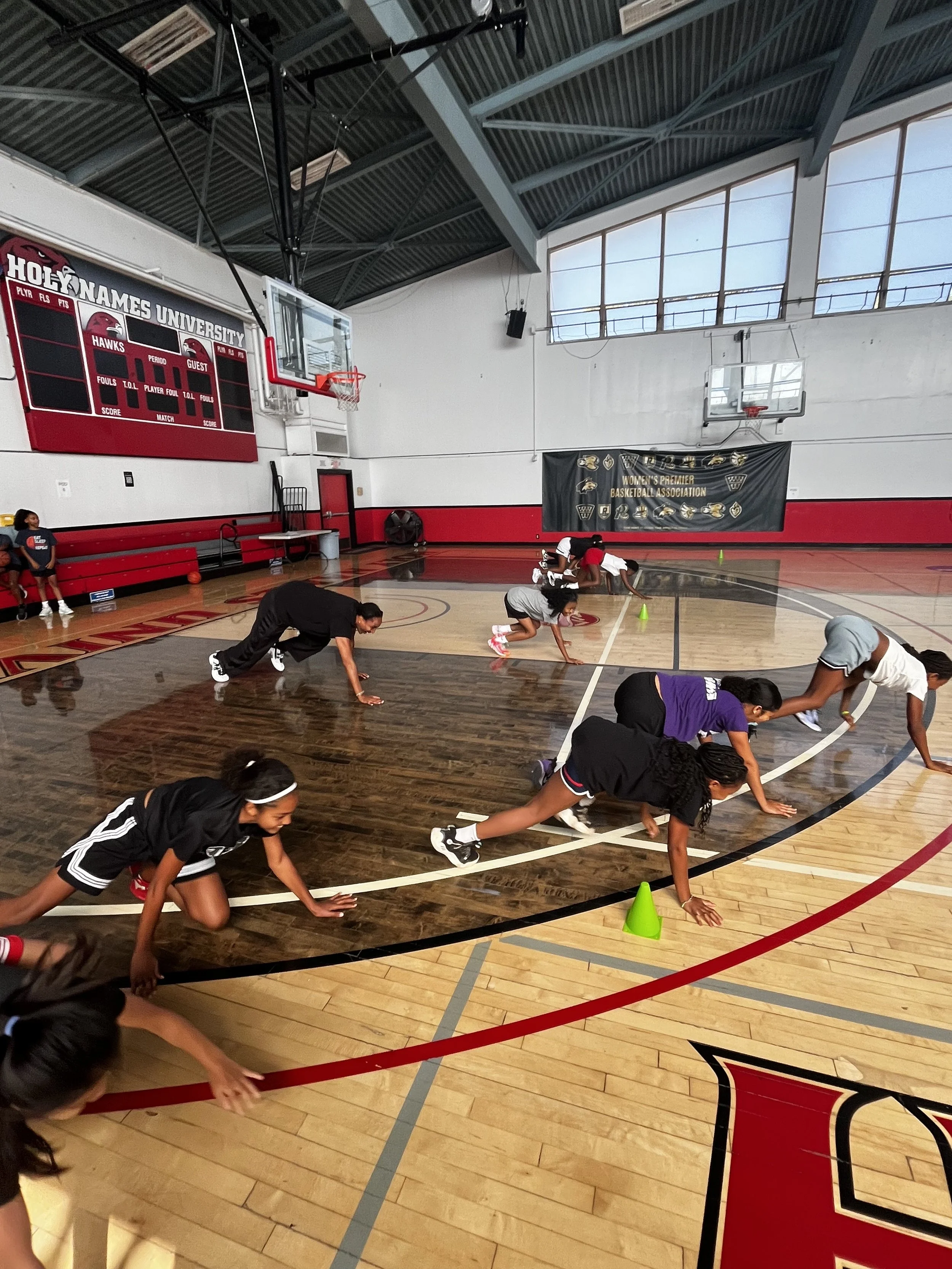 Group of young girls doing physical training exercises on a basketball court indoors, with a basketball hoop and scoreboard in the background.
