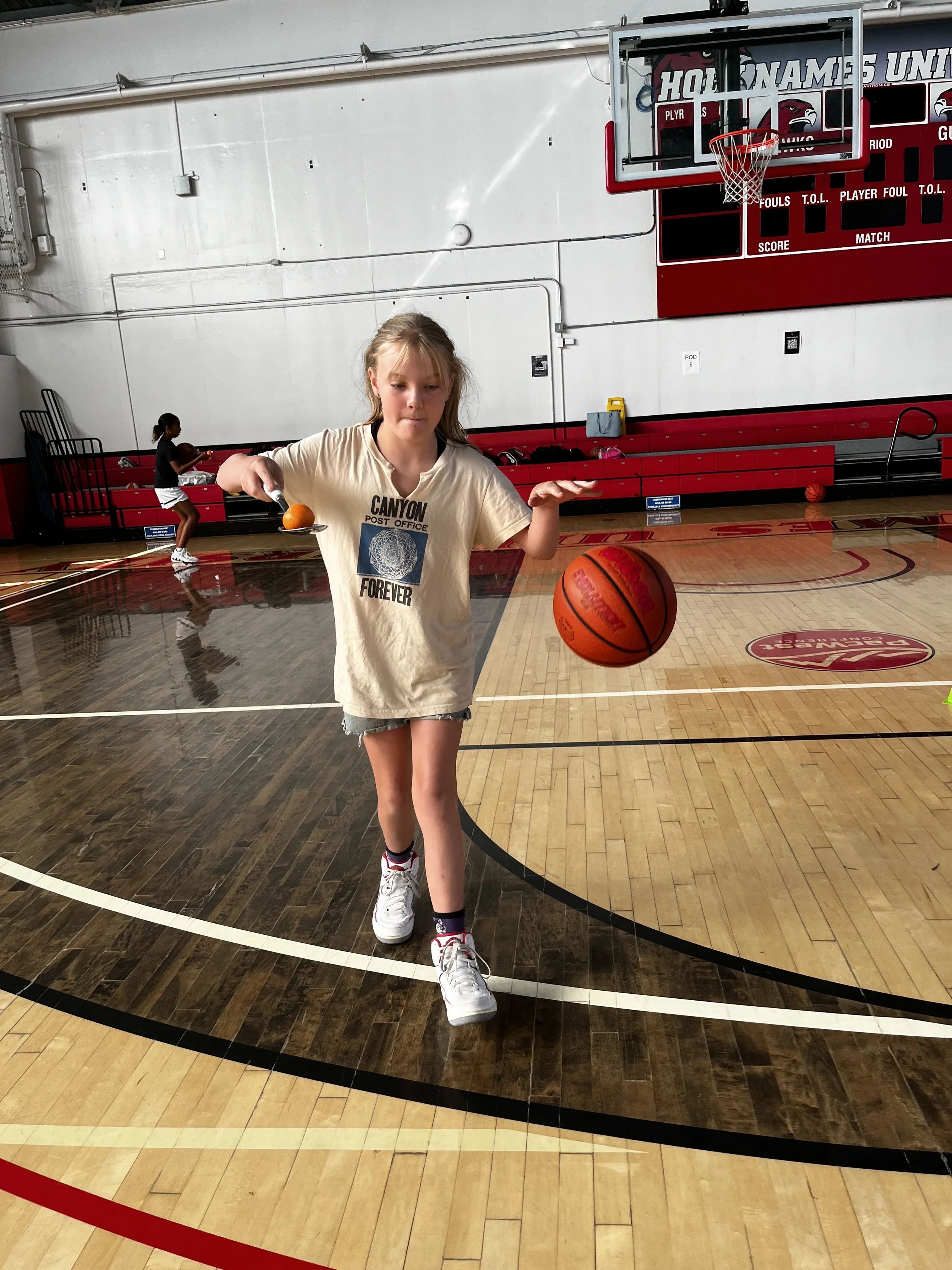 A young girl playing basketball on an indoor court with another girl in the background. The girl in front is holding a basketball with one hand and a tennis ball with the other, while walking on the court.