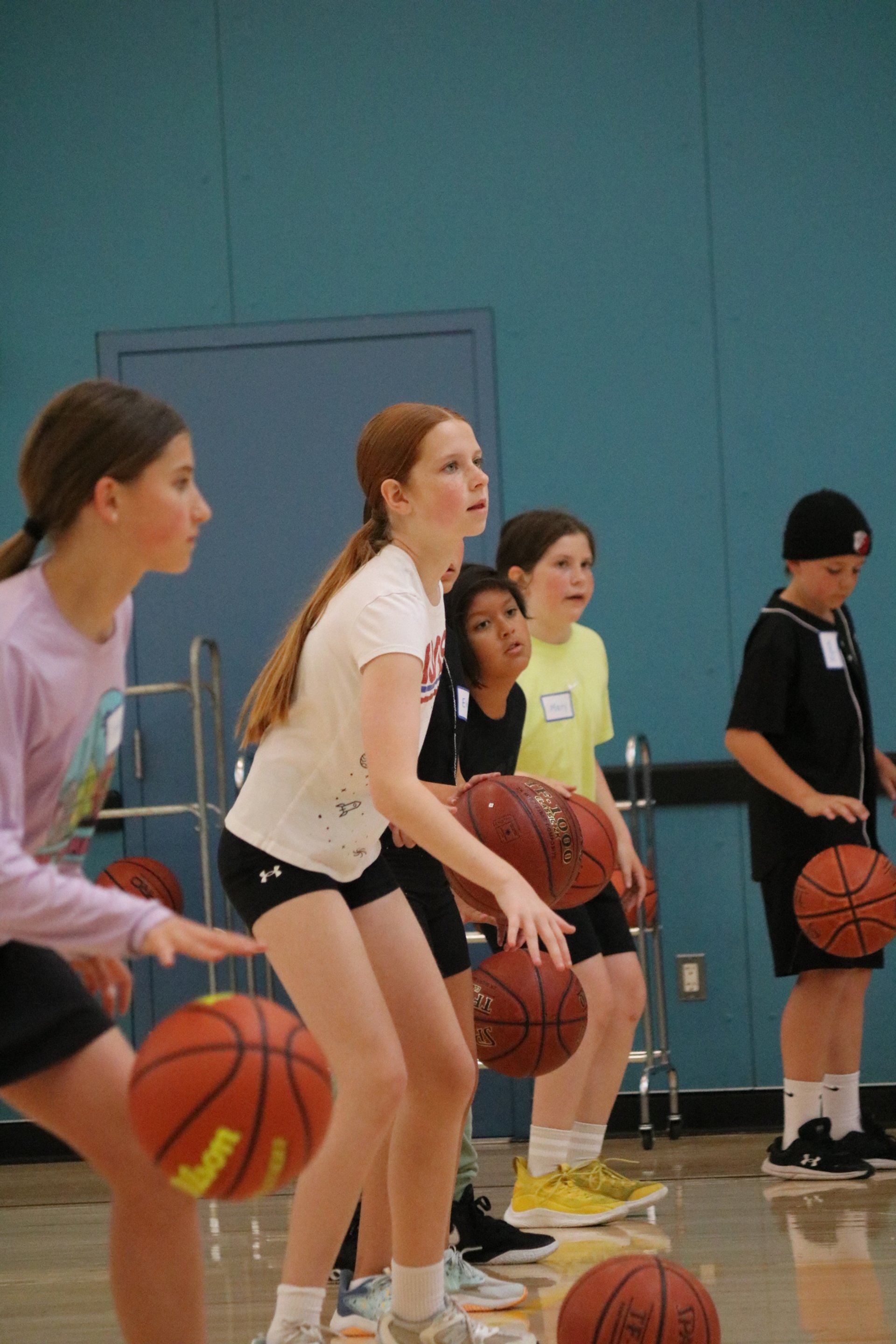 Young girls practicing basketball in a gym, holding basketballs, and standing in a row.