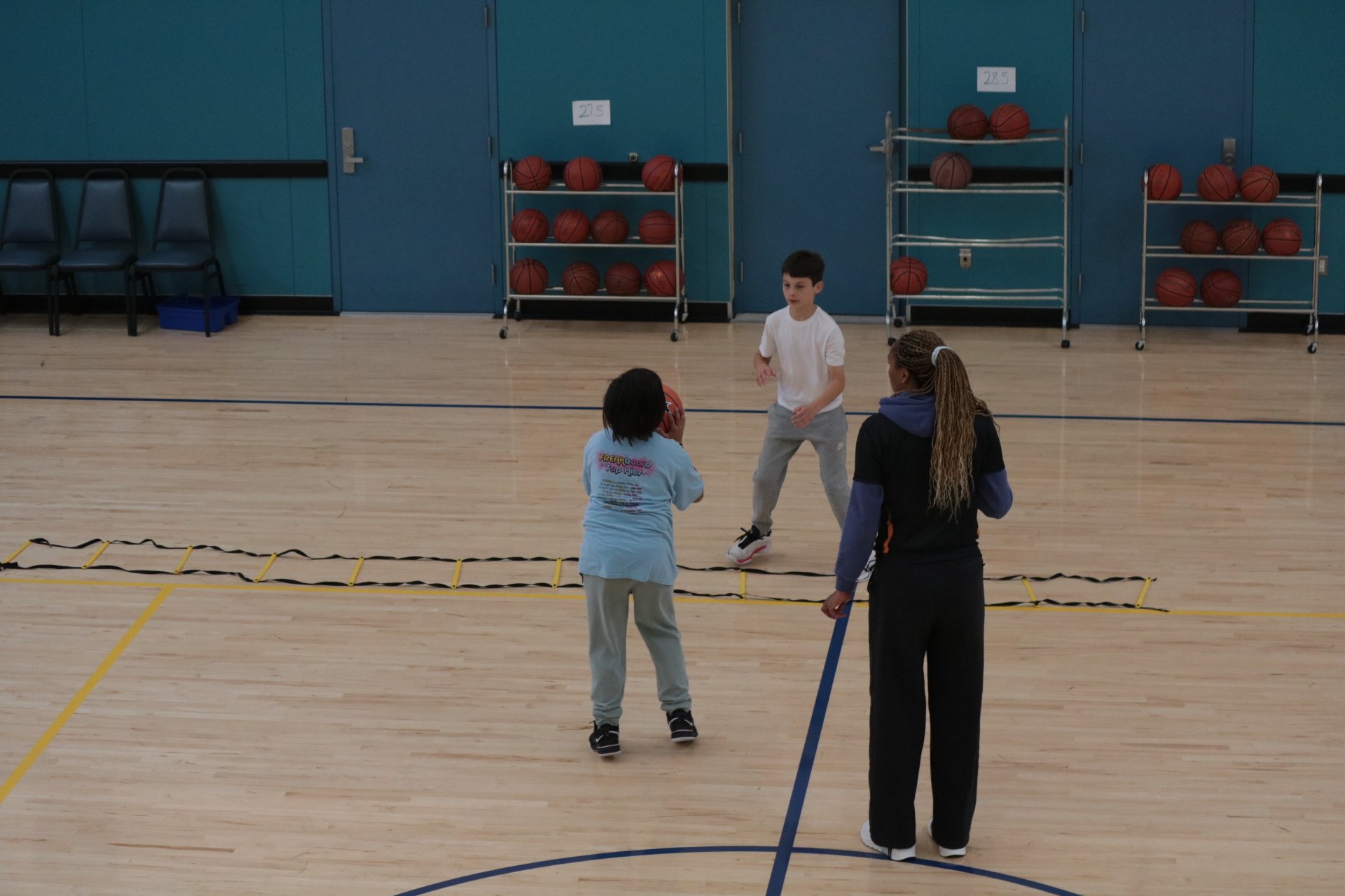Children and a coach in a gym practicing basketball, with racks of basketballs in the background.