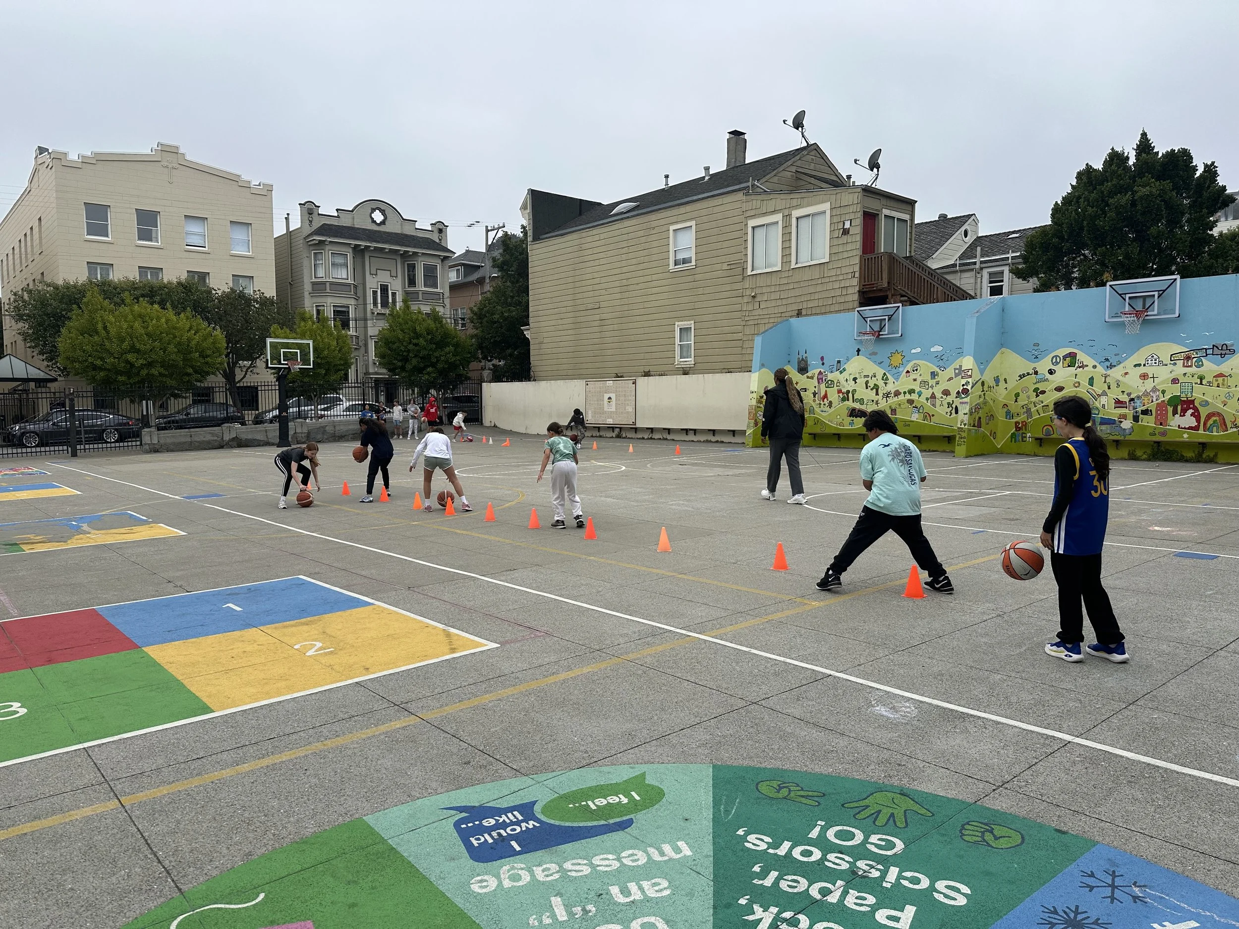 Children practicing basketball drills on an outdoor court under adult supervision, with orange cones set up for drills, in a city neighborhood.