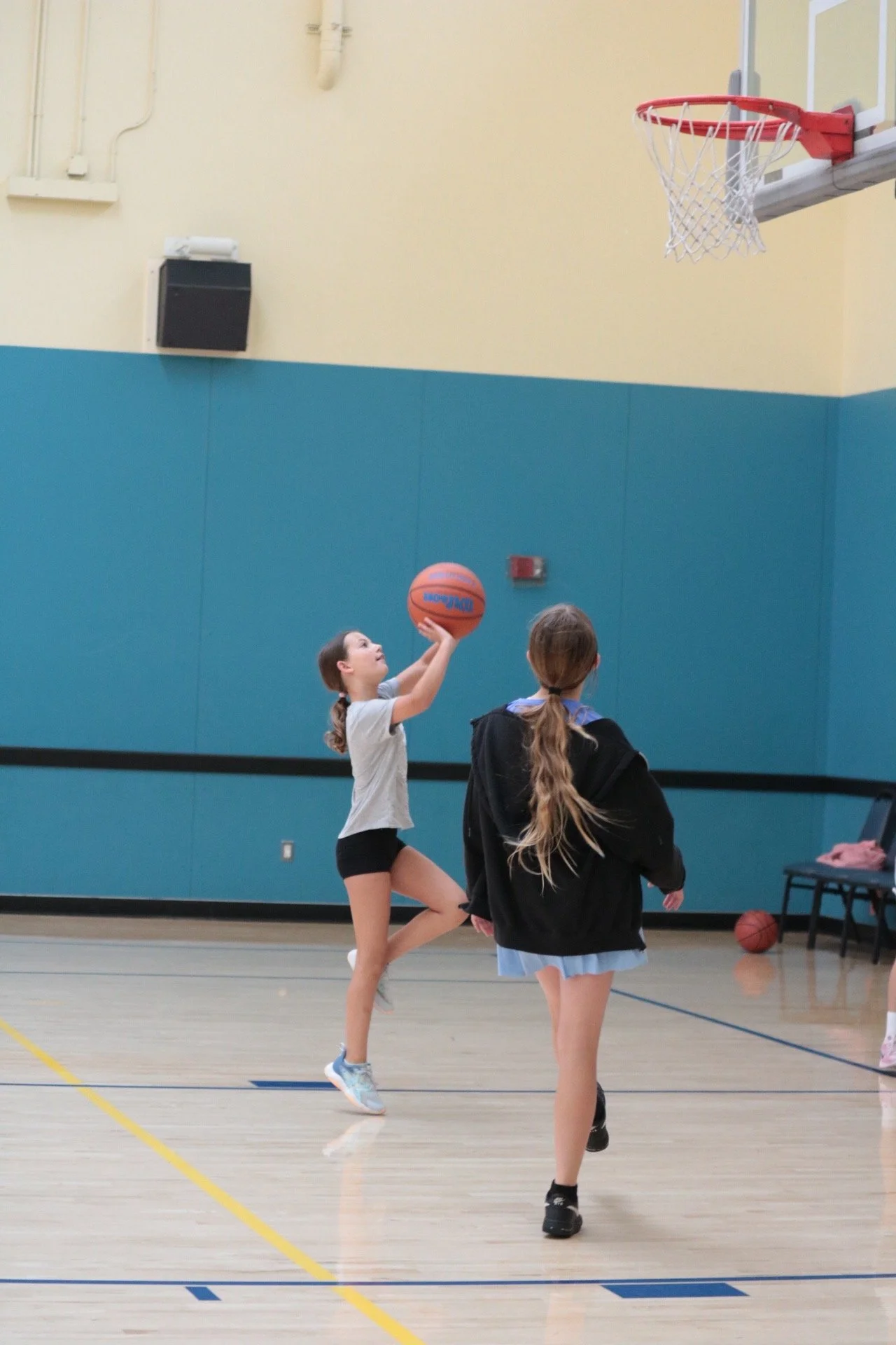 Young girl playing basketball in an indoor gym, about to shoot toward the hoop while another girl watches.