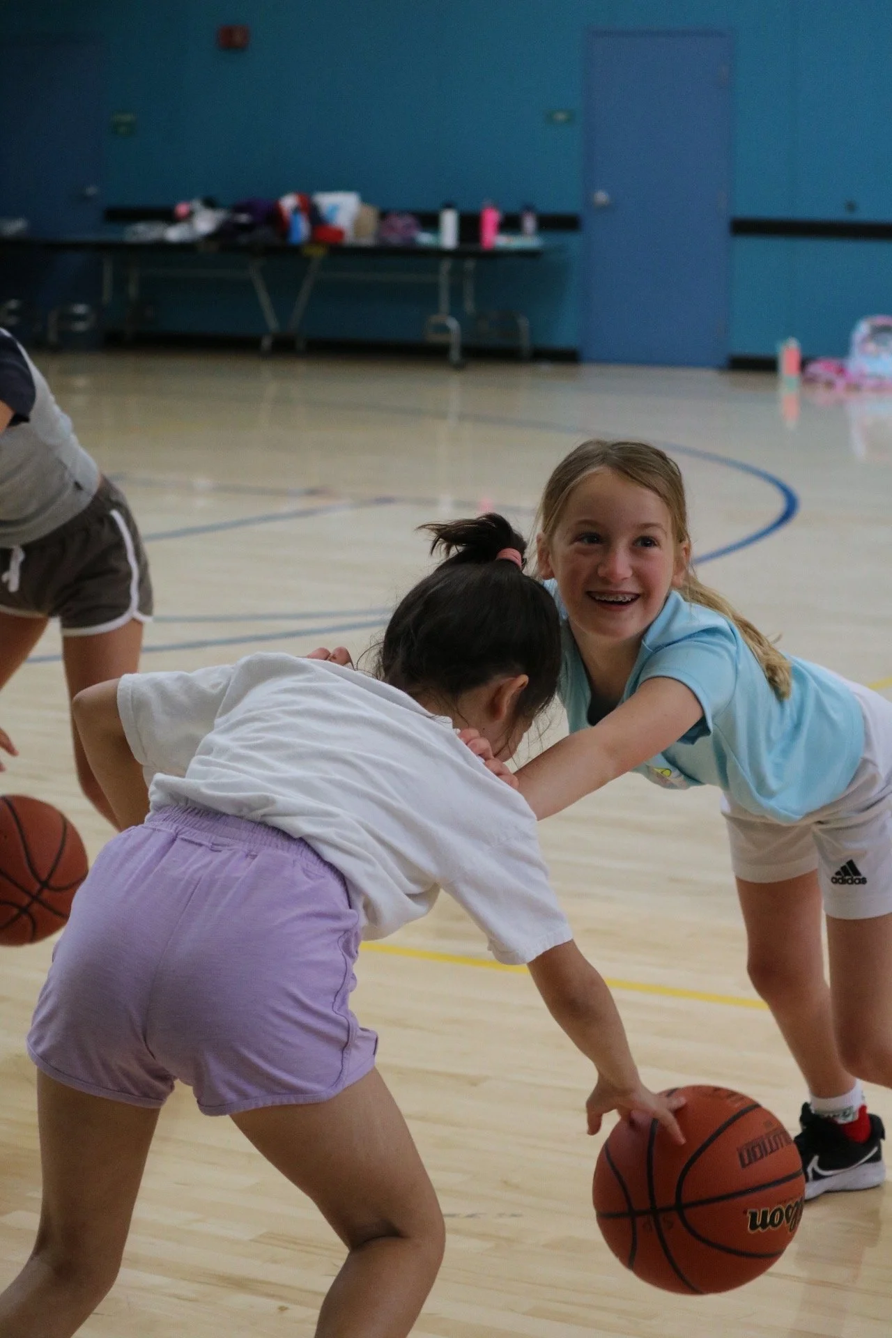 Two young girls playing basketball in a gym, with one girl in a white shirt and purple shorts reaching for the basketball while the other girl in a blue shirt and white shorts is smiling.