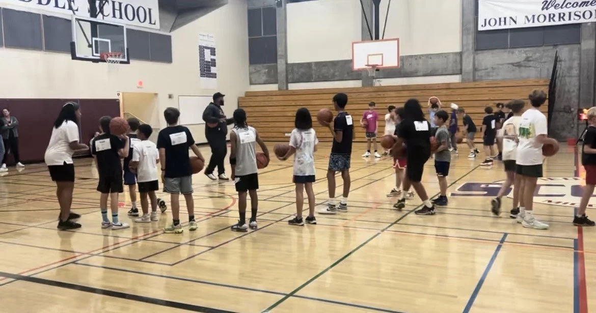 Children in a gymnasium participating in a basketball practice or class, with coaches or instructors instructing.