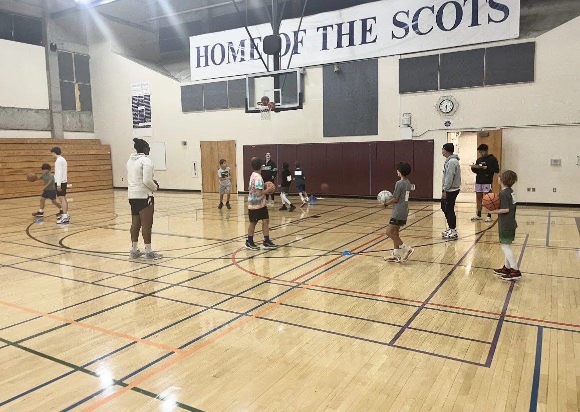 Children and adults playing basketball in a gymnasium with a large banner reading 'HOME OF THE SCOTS' hanging above the court.