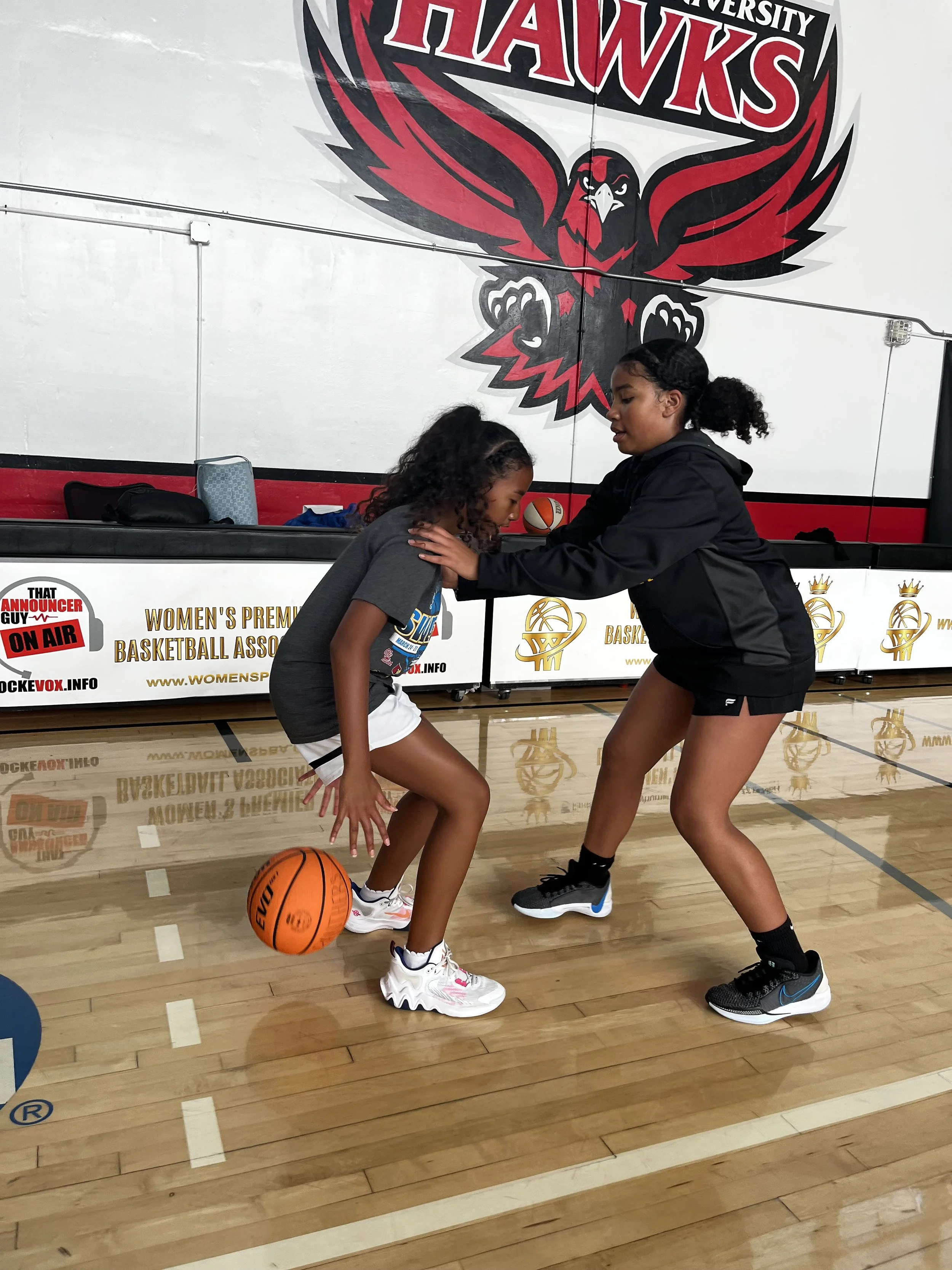 Two young women practicing basketball on an indoor court. One is bending over with a basketball near her foot, while the other is leaning forward and holding her shoulders. A large red and black hawk logo and basketball-related banners are visible on the wall behind them.