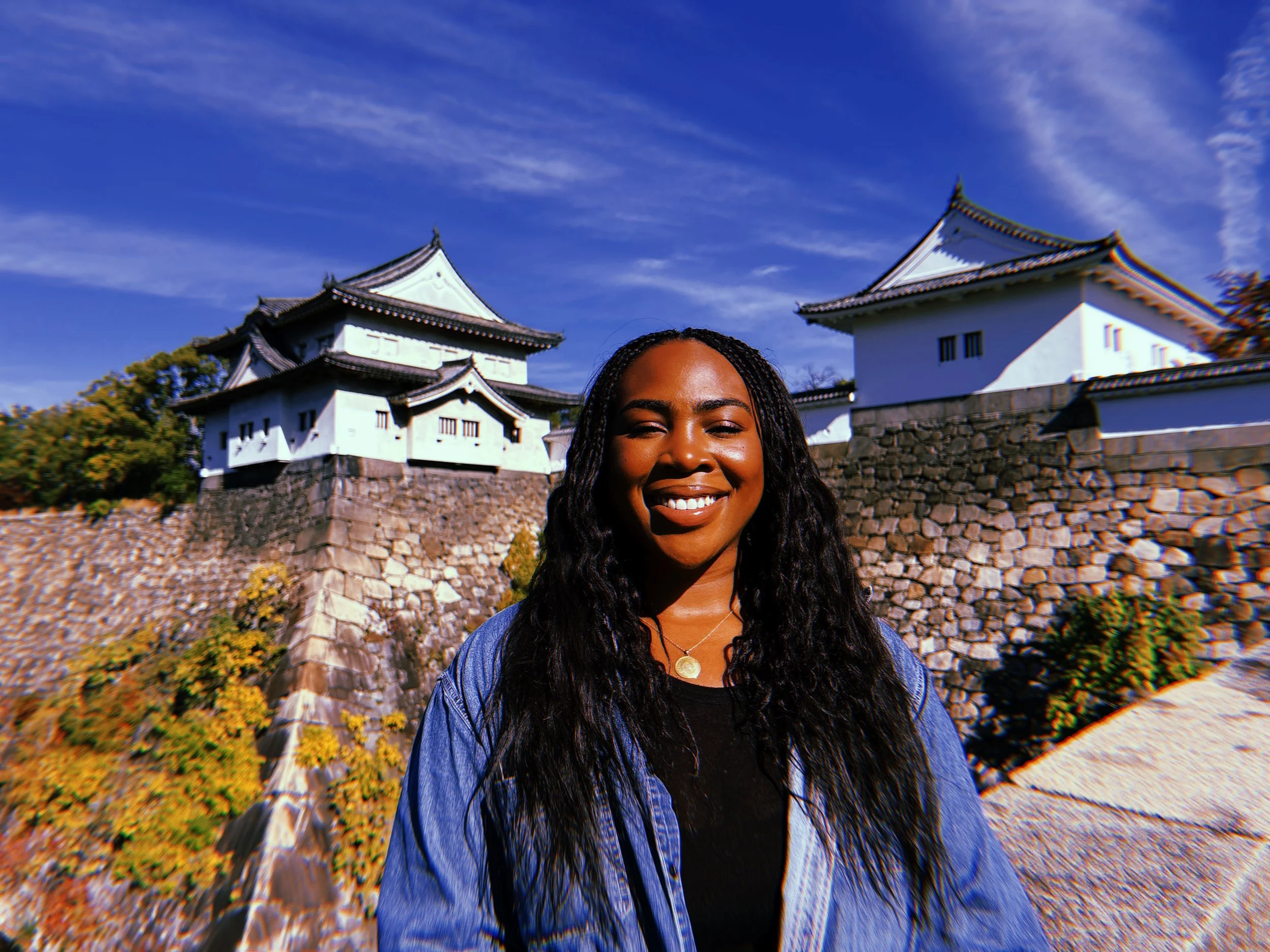 A smiling woman with long black curly hair standing outdoors in front of a traditional Japanese castle with white walls and a stone foundation, under a blue sky.