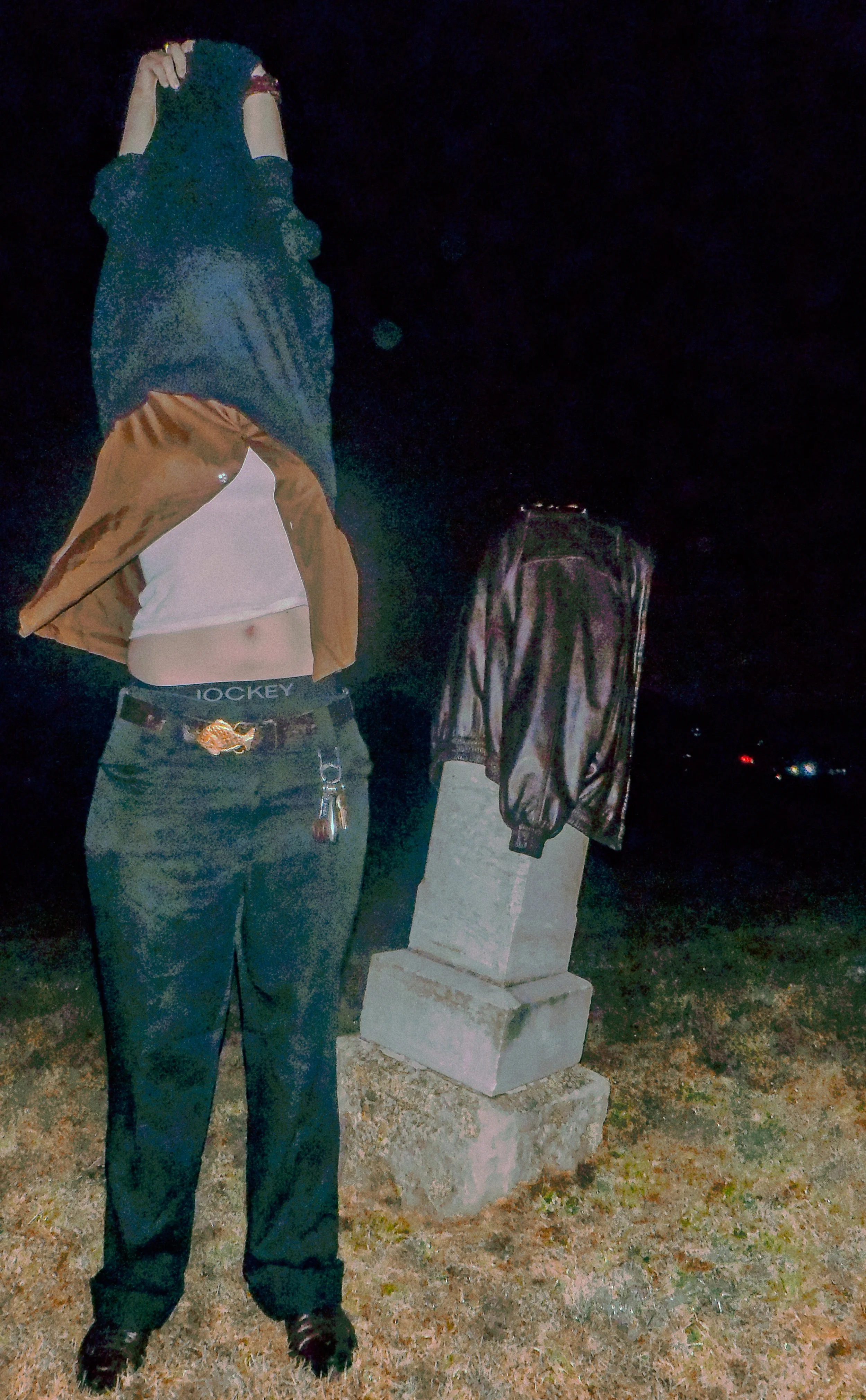A person standing outdoors at night with their hoodie up, wearing dark pants, a white shirt, and a brown jacket, with keys hanging from their pocket, and a statue with a black jacket on a concrete base in the background.