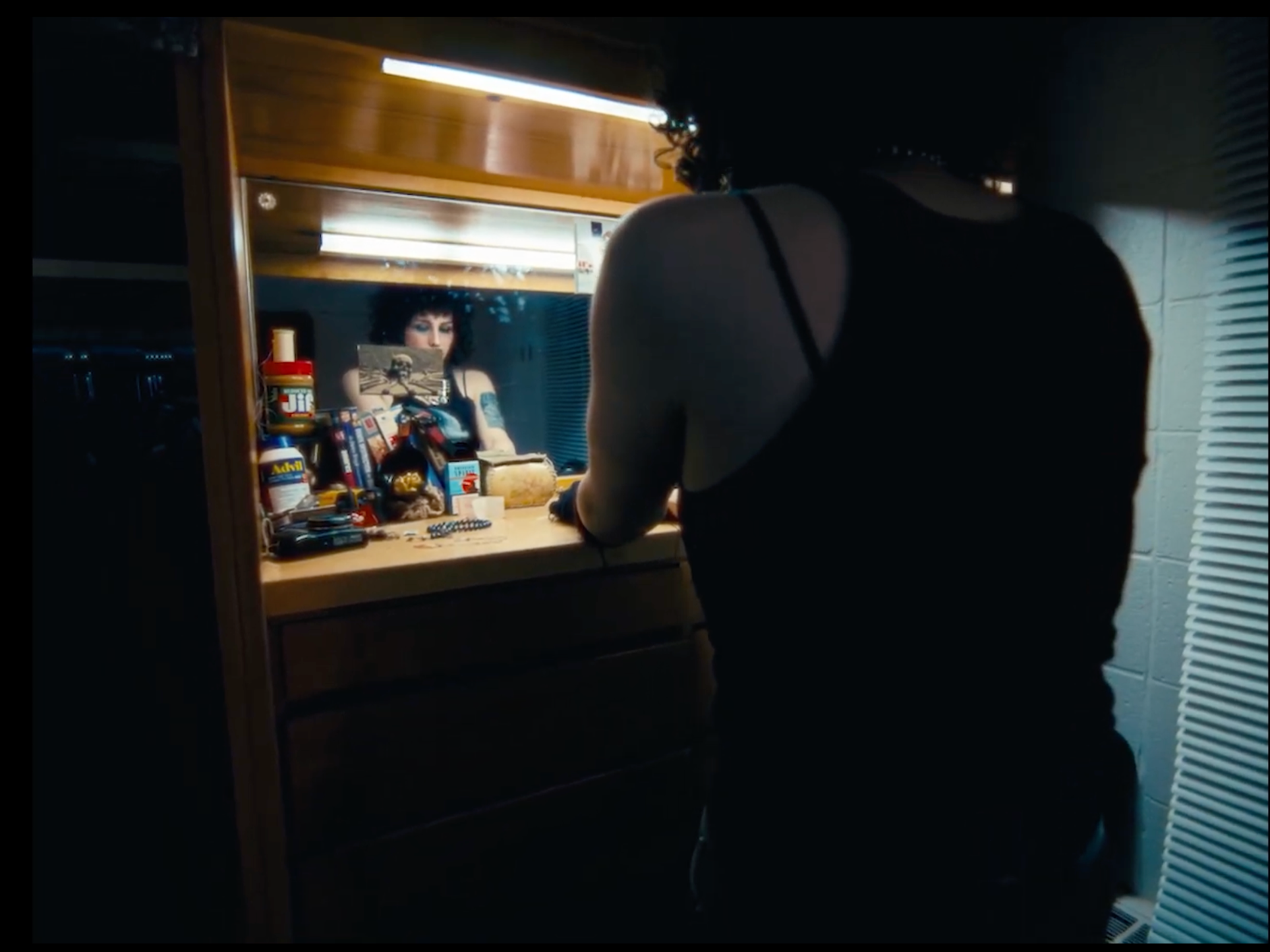 A woman with curly hair in a black tank top looking into a mirror at a cluttered dresser in a dimly lit room.
