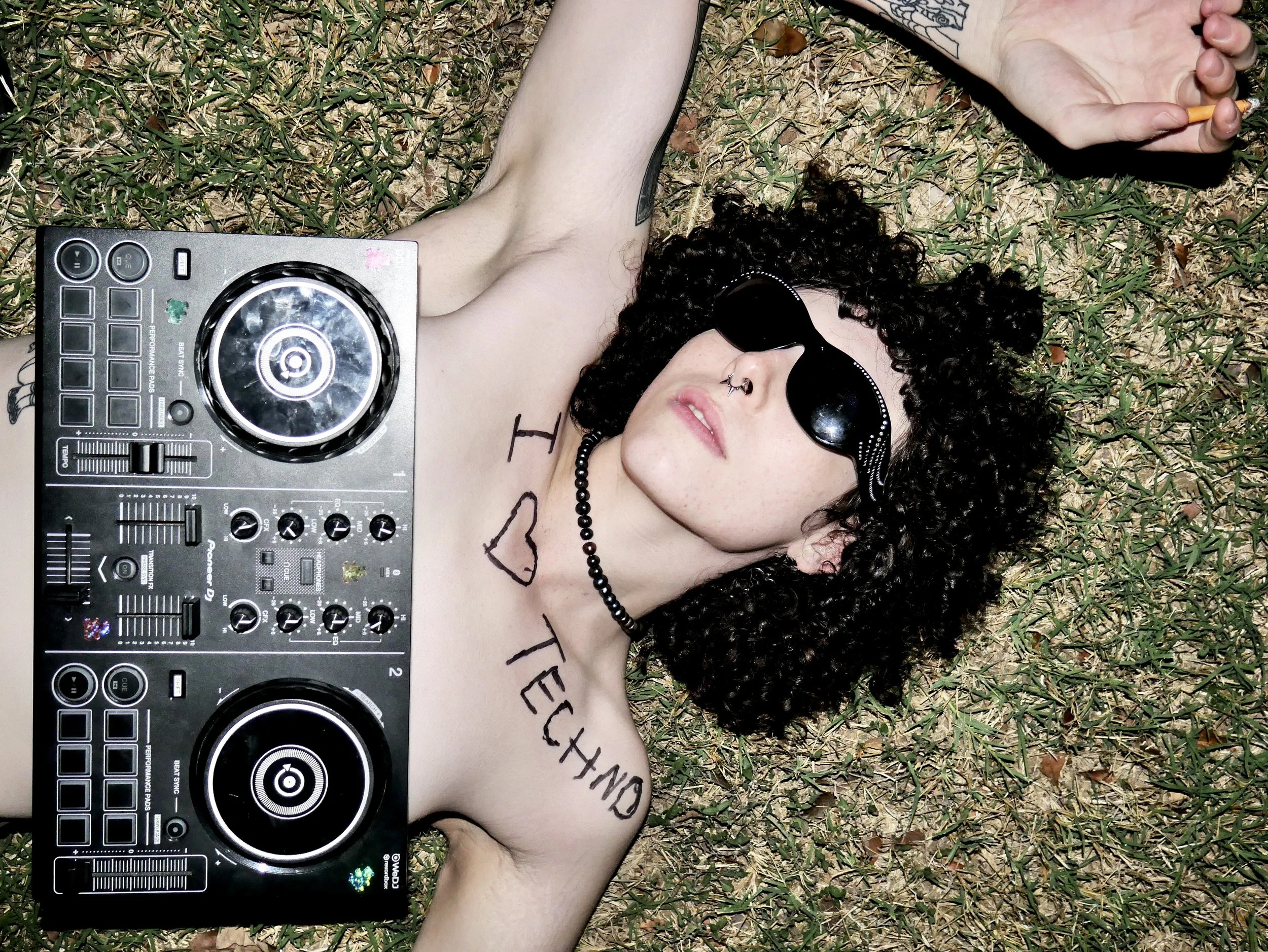 A woman with curly hair is lying shirtless on grass, wearing sunglasses, a choker, and a nose ring, holding a cigarette in her right hand. The words "I ♥ TETNO" are written on her chest.