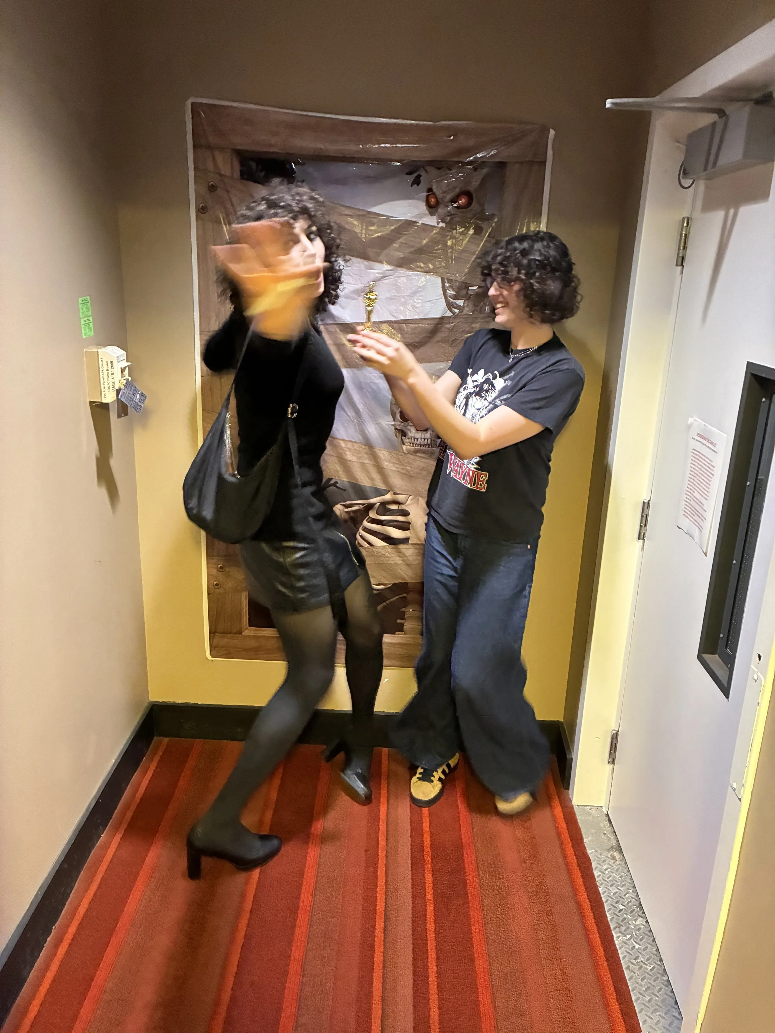 Two women with curly hair, one wearing black with high heels and the other in a black t-shirt and baggy pants, exchanging a small object in a hallway with Halloween decorations in the background.