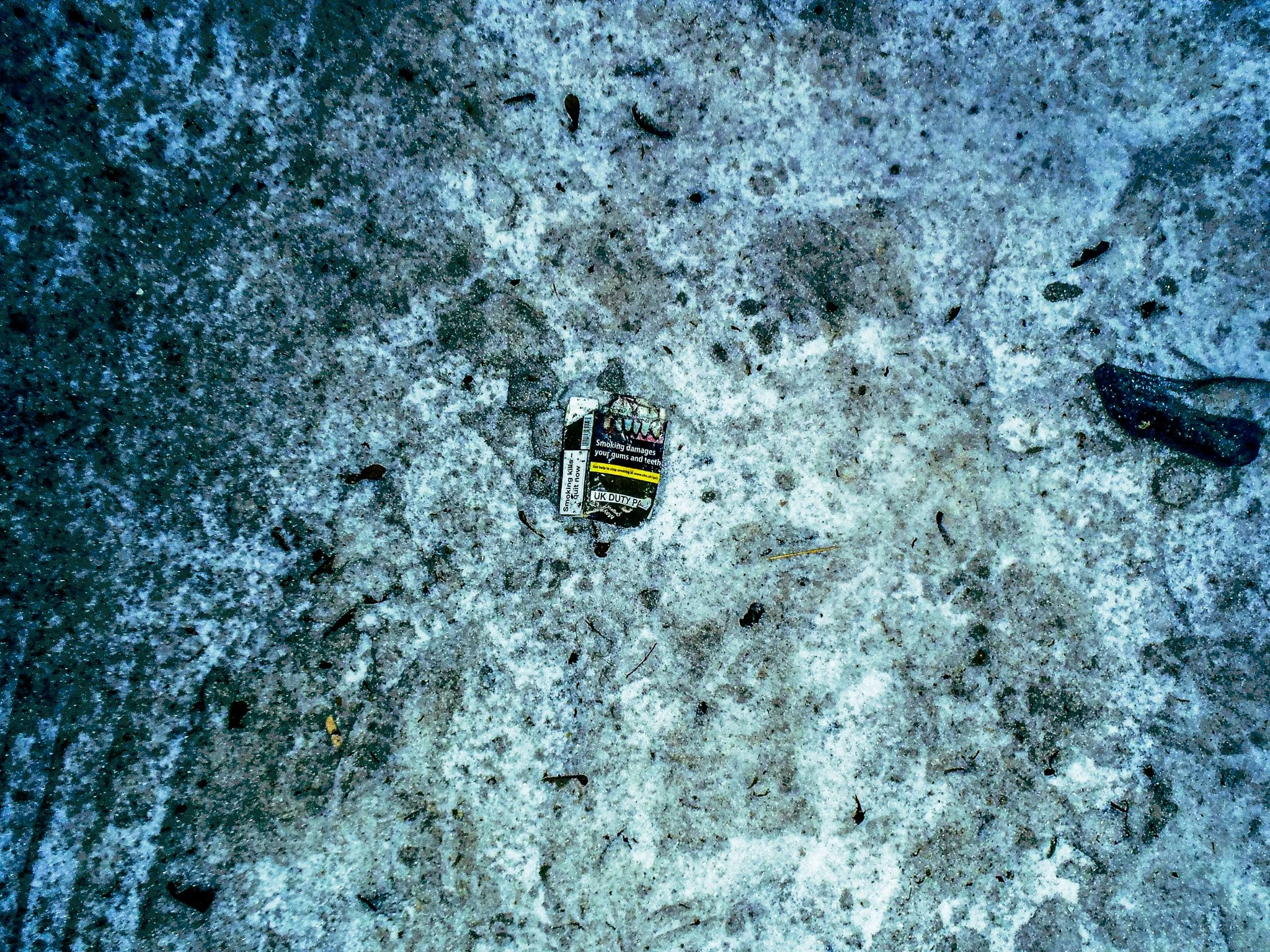 A crushed cigarette pack lying on a dirty, snowy pavement with small debris and dark spots.