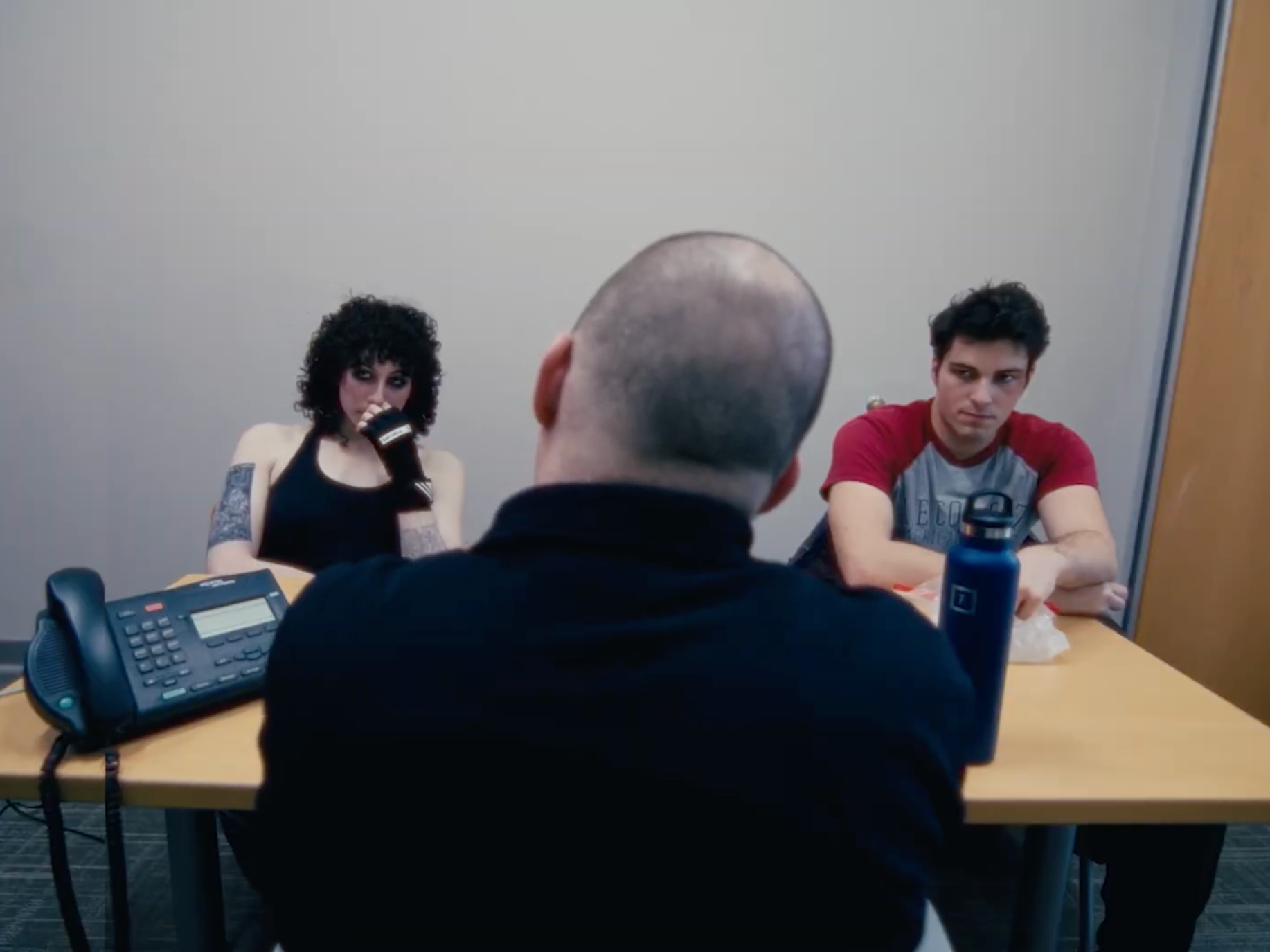 Two young adults, a woman and a man, are seated at a table across from a man with a shaved head, in a conversation. The woman has curly dark hair and tattoos, and is wearing black gloves and a black tank top. There is a landline phone and a water bottle on the table. The setting appears to be an office meeting room.