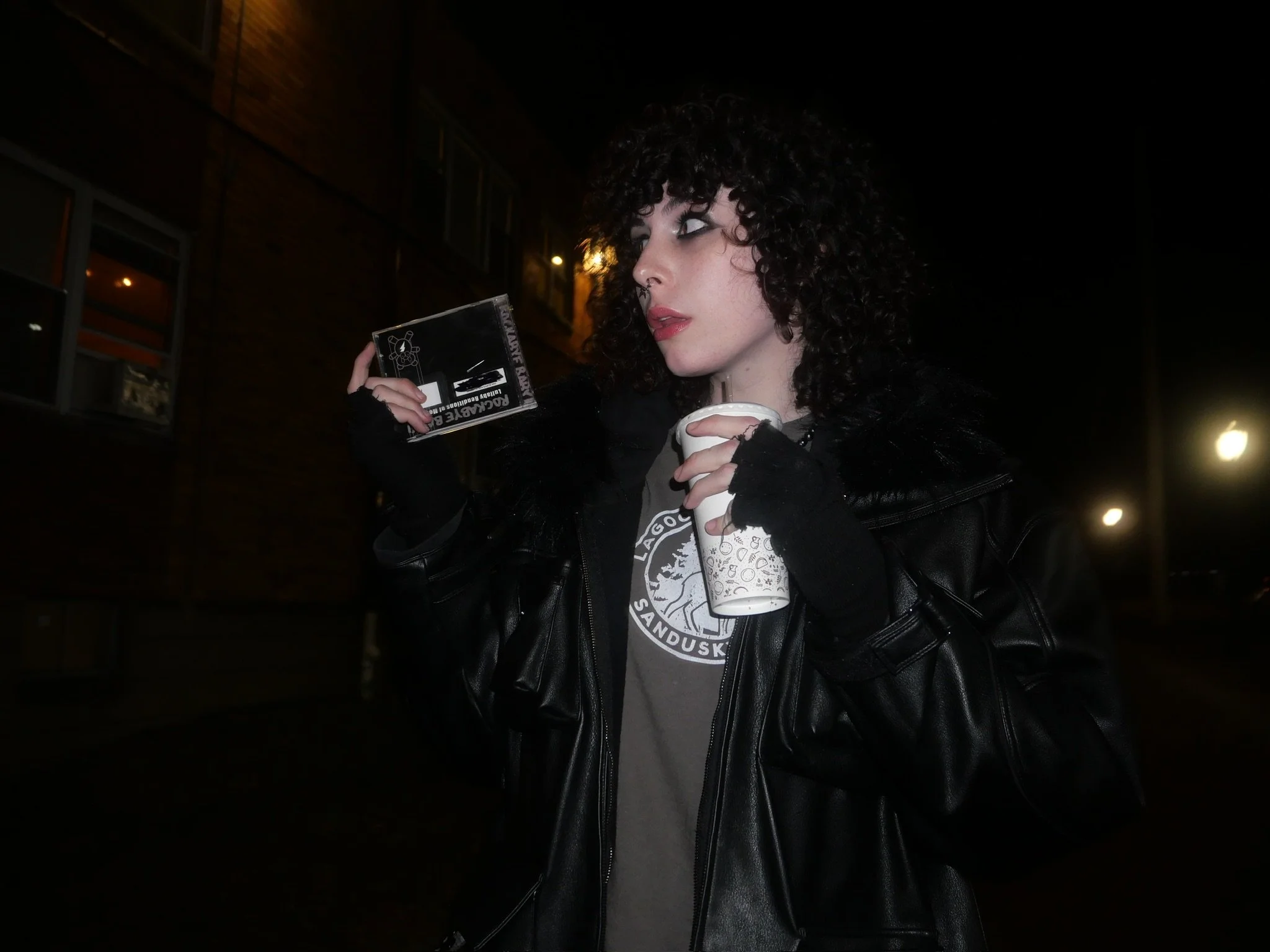 A woman with curly dark hair, wearing black gloves, a graphic t-shirt, and a black leather jacket, stands outdoors at night, holding a drink in a paper cup and a small packet or box, with a building and streetlights in the background.