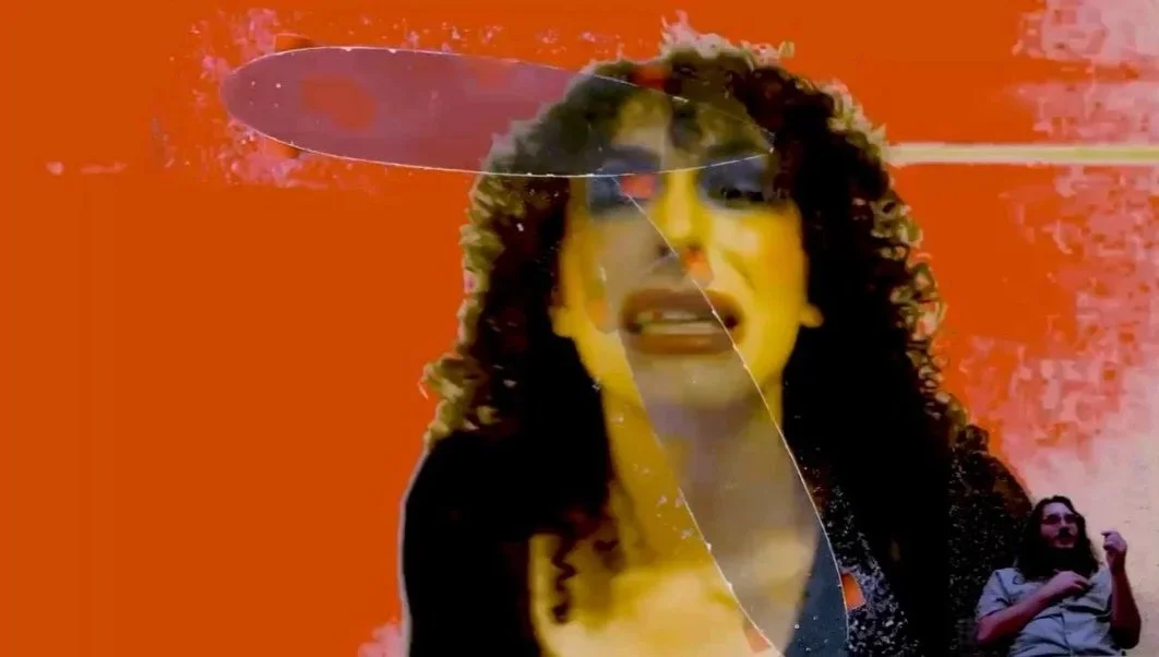 A woman with curly hair and makeup standing against an orange background, with a glass panel and a person sitting in the corner.