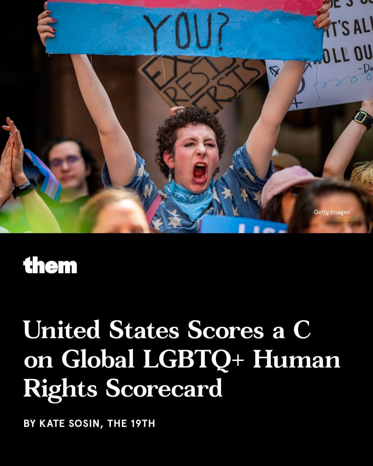 A person with short curly hair and a blue bandana, passionately shouting and holding a sign that says 'YOU?'. Several other people are holding signs and clapping at a protest or rally.
