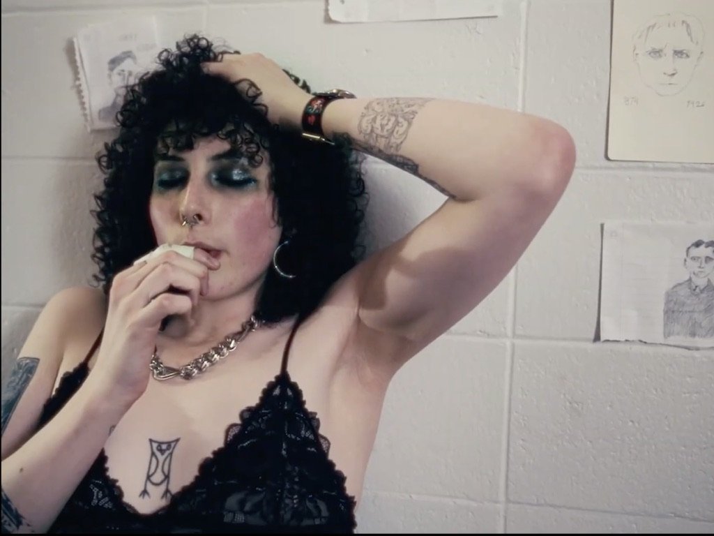 A woman with dark curly hair, heavy makeup, septum piercing, and tattoos, wearing a black lace top, touching her head with one hand. She is standing against a white brick wall with sketches taped to it.