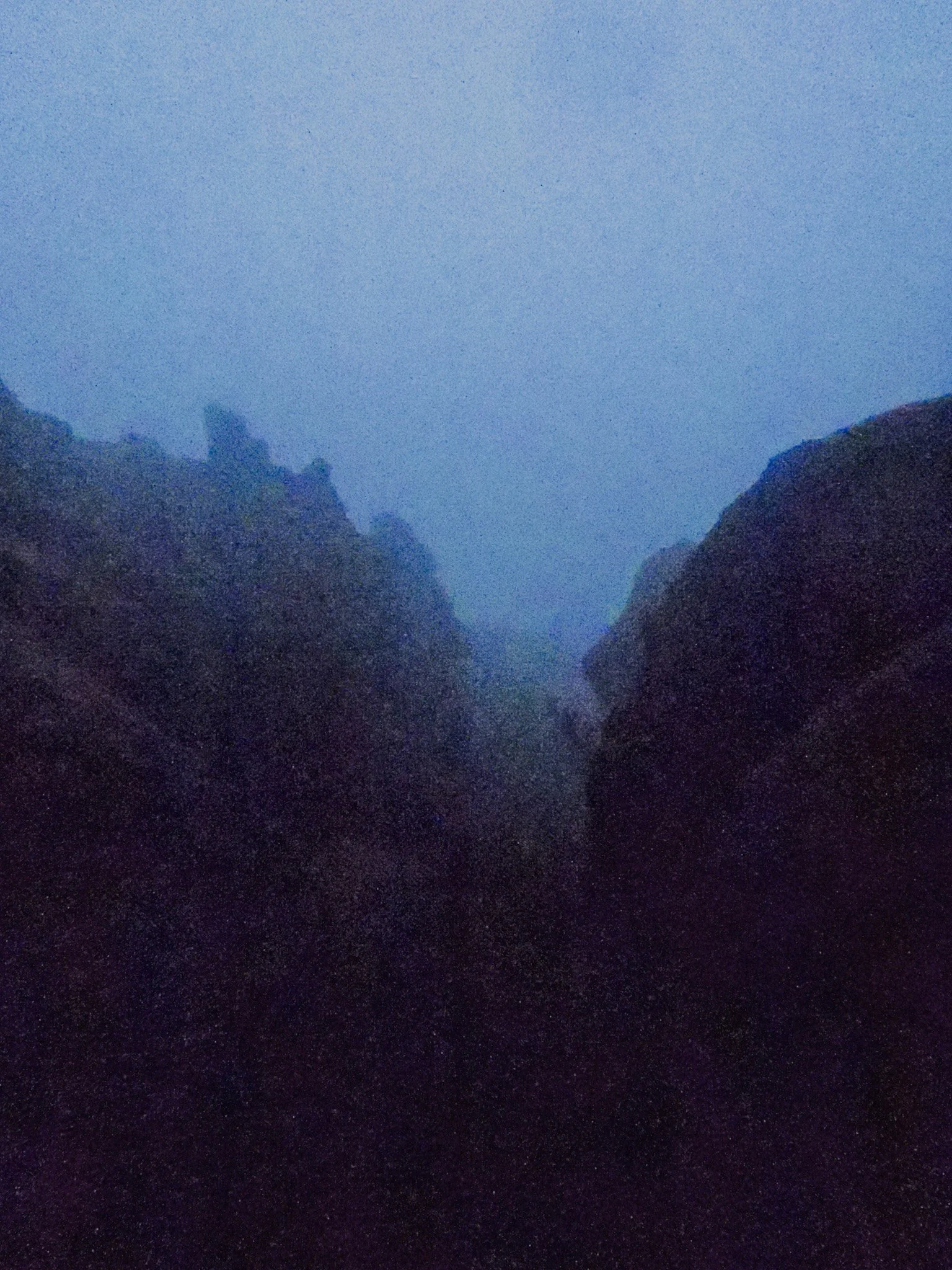 A foggy mountain canyon with steep rocky cliffs on both sides and a narrow, dark passageway between them