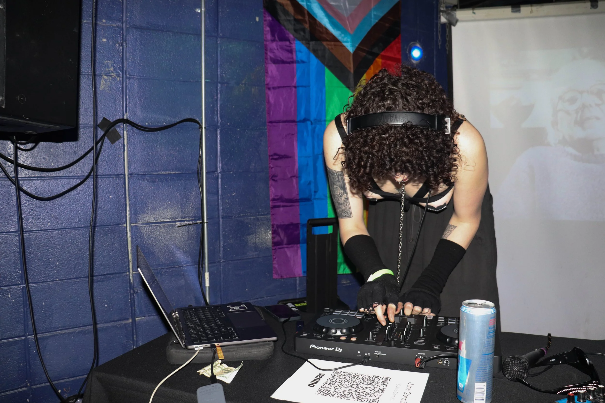 A DJ with curly hair wearing headphones, a black sleeveless outfit, and fingerless gloves is mixing music at a DJ booth. There is a laptop, a can of Red Bull, a microphone, and a QR code on the table. The background features a colorful flag and a projected image of a person with glasses.