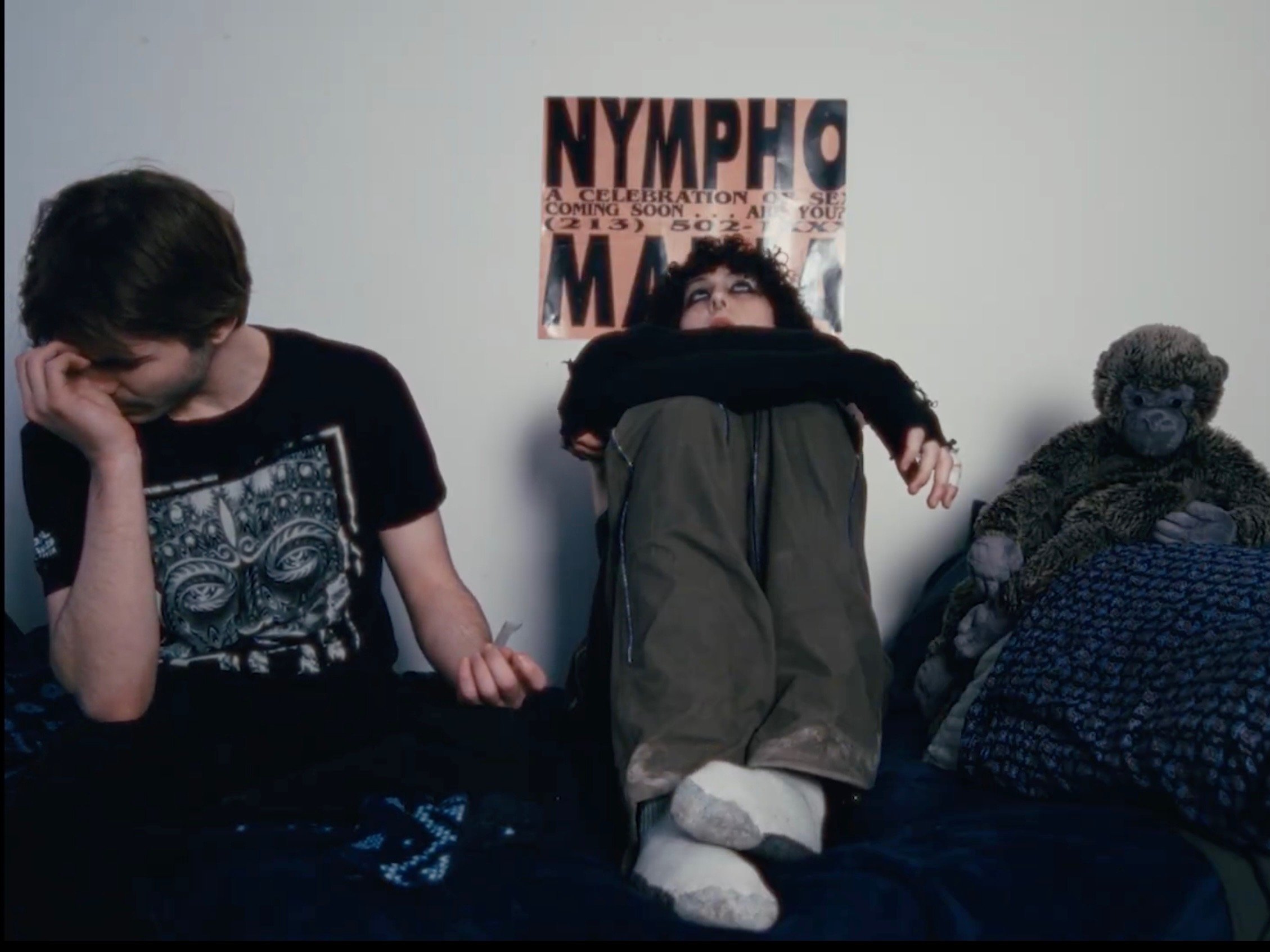 people sitting on a bed with a poster on the wall behind them. The person on the left has short dark hair and is wearing a black T-shirt with a graphic design, looking down and touching their forehead. The person in the middle has curly hair, wearing dark clothing, sitting with their legs crossed, and looking up. The person on the right is a rain forest animal plush toy resembling a monkey, sitting upright and holding its legs.
