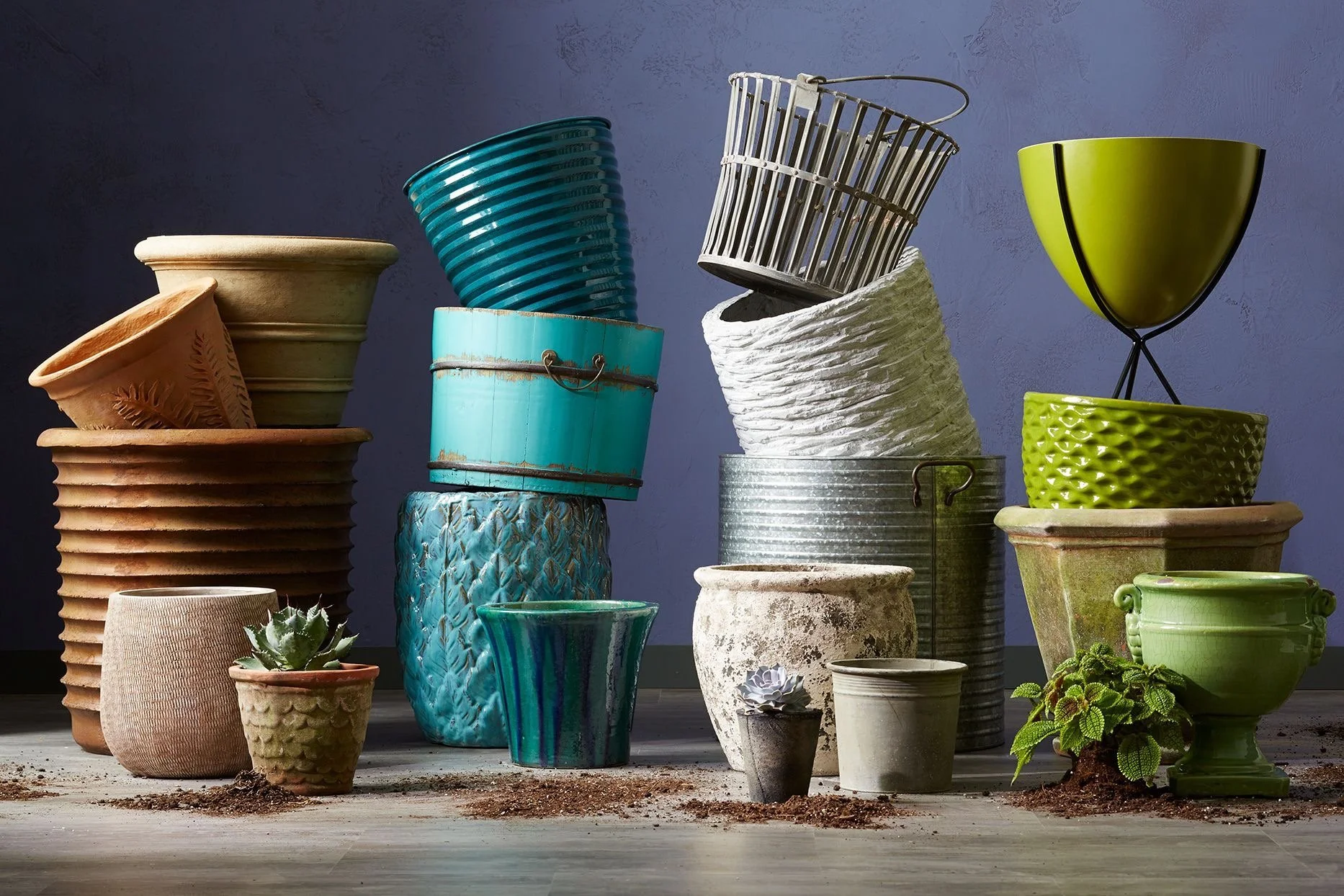 Stacked and scattered flower pots and planters of various sizes, colors, and textures on a wooden surface with soil and a purple wall in the background.