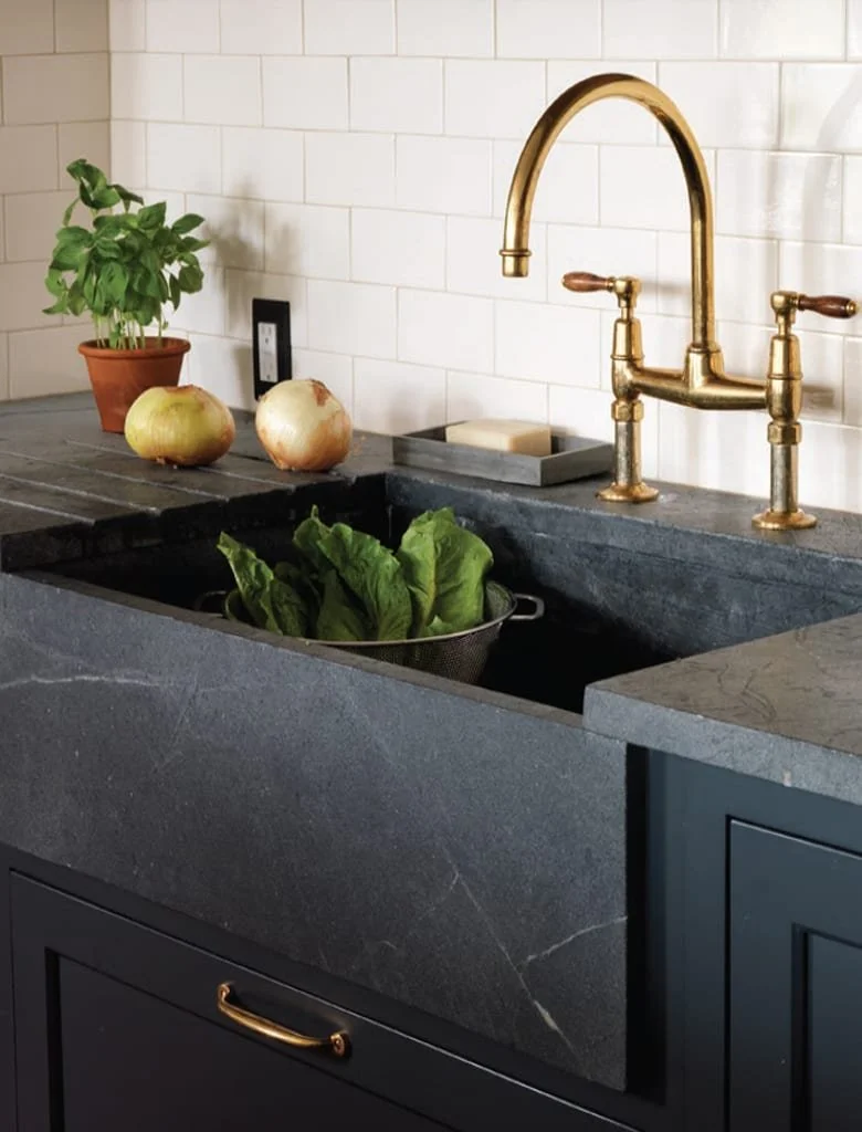 Kitchen sink with brass faucet, a potted basil plant, two onions, and leafy greens in a colander.