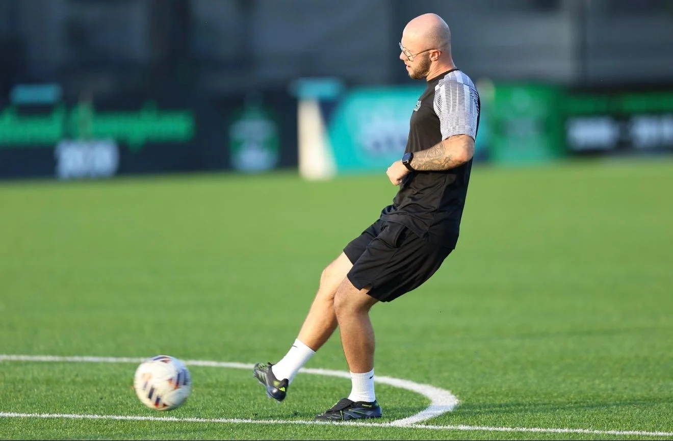 Man with a bald head, glasses, and tattooed arm wearing black athletic shirt and shorts playing soccer on a field.