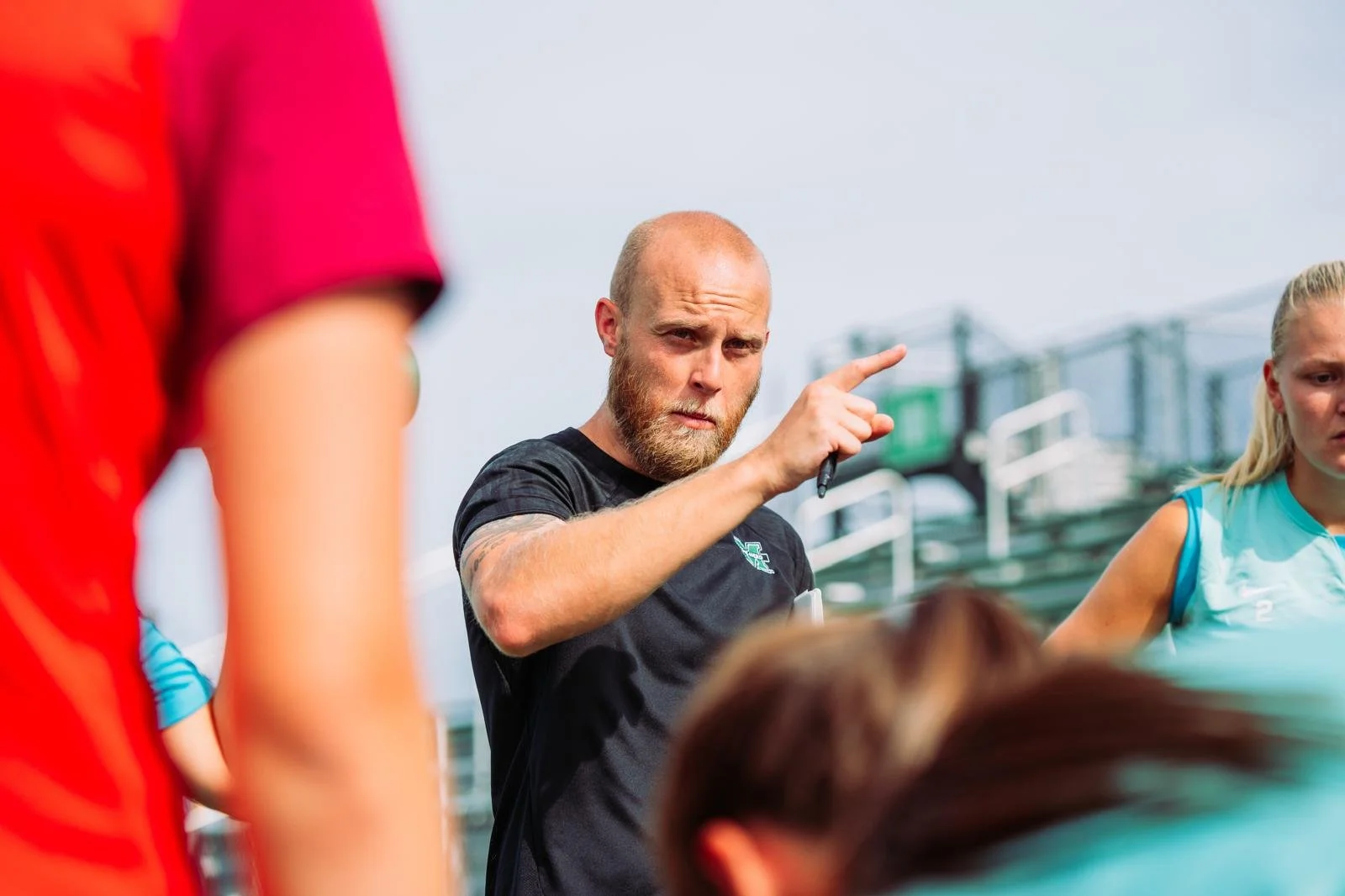 A male coach with a beard, dressed in black, gesturing with his finger during an outdoor sports practice with athletes around him.