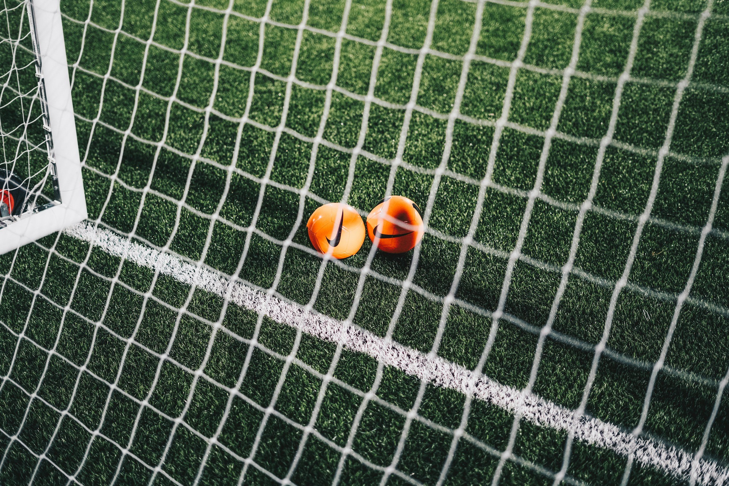 Two orange soccer balls with Nike swoosh logos behind a soccer net, near the goal line on a green artificial turf field.