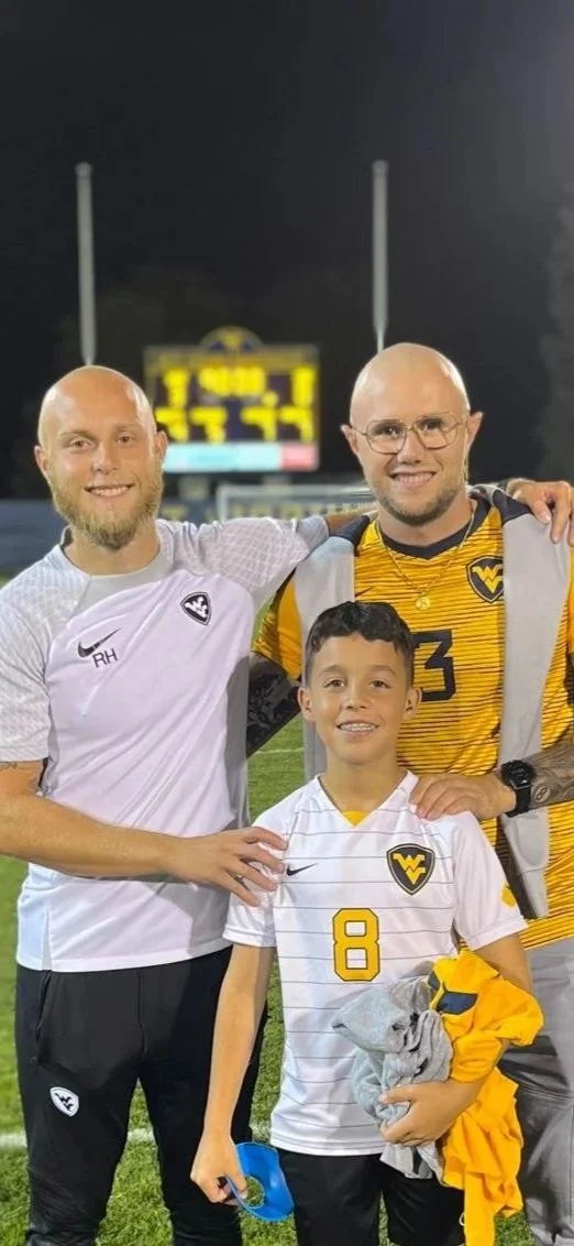 Three people, including a young boy, standing on a football field at night, all wearing West Virginia University football gear, smiling for a photo.